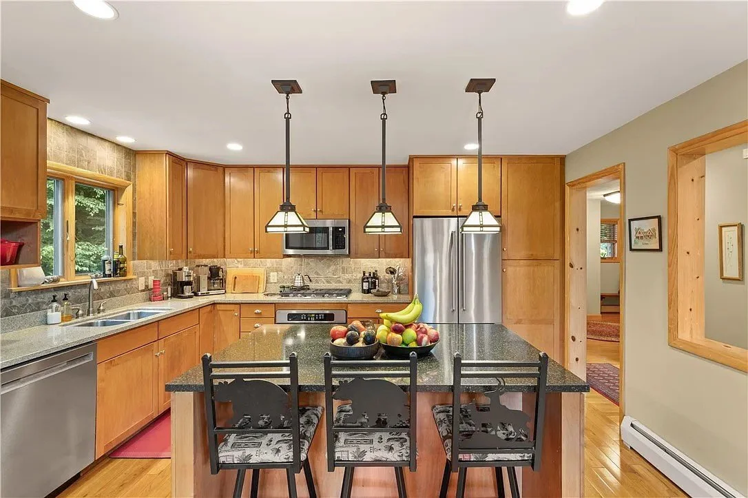 A modern kitchen with wooden cabinets, a black granite island, stainless steel appliances, and three pendant lights over the island. There are bowls of fruit on the island and a window showing greenery outside.