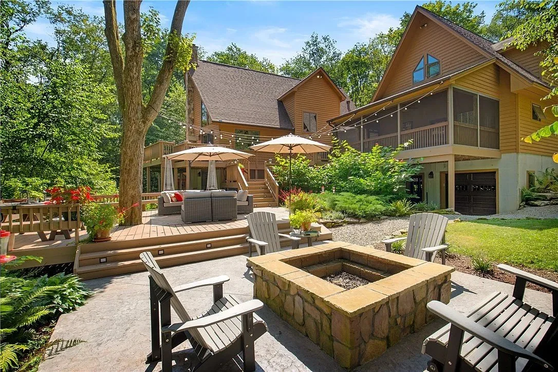 Backyard patio area with stone fire pit surrounded by four Adirondack chairs, with a wooden deck, chairs, umbrellas, and a house in the background, surrounded by trees and greenery.