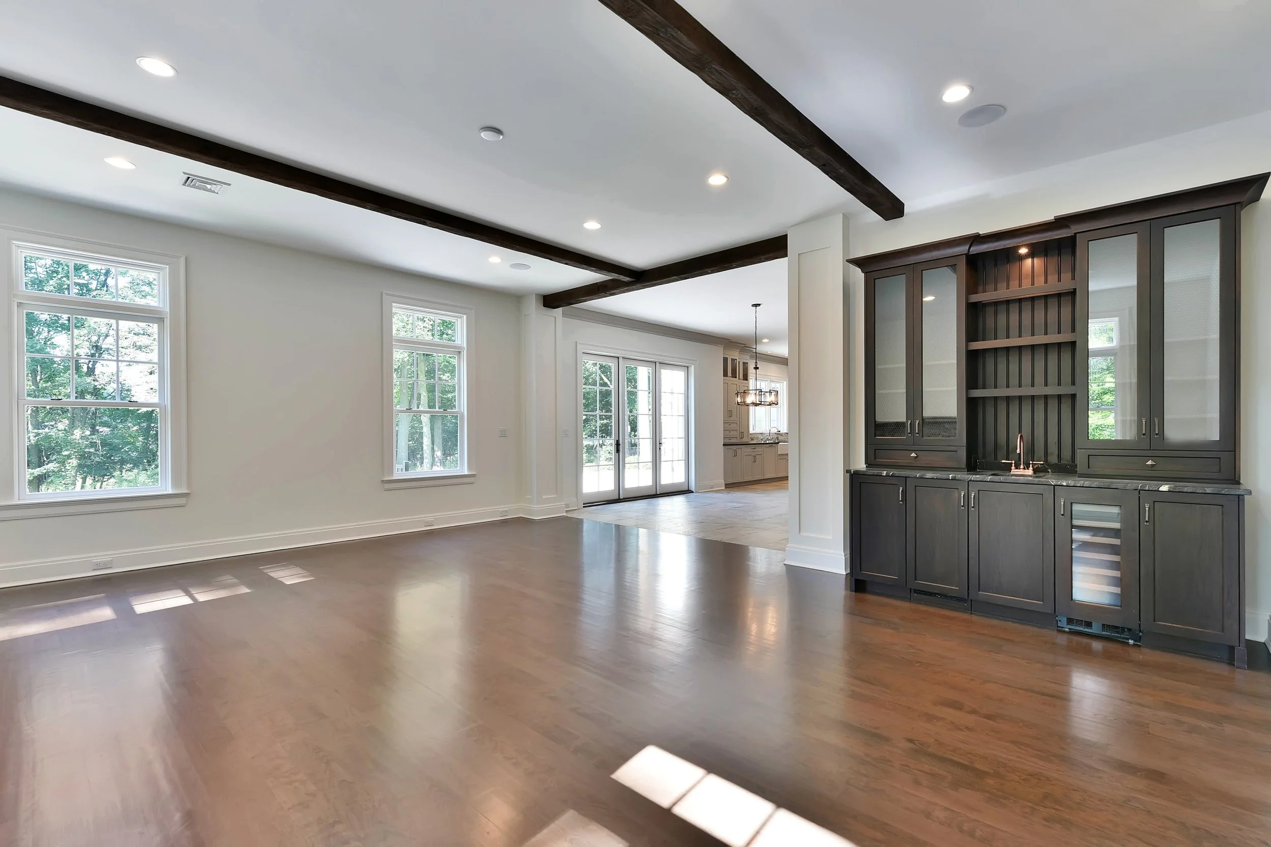 Empty living room with hardwood floors, white walls, large windows, and a dark wood built-in bar area.