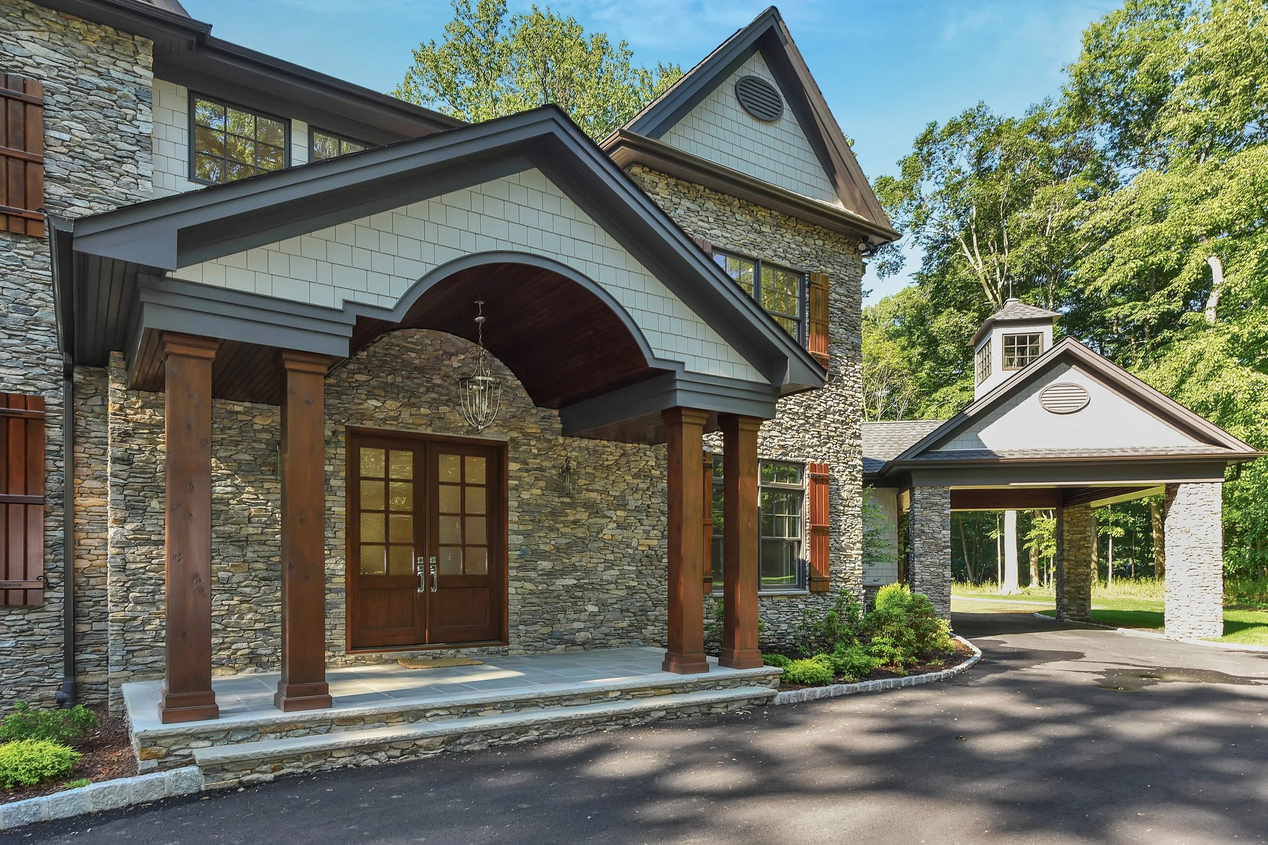 Front view of a house with stone and shingle exterior, wooden columns, and a driveway, surrounded by trees.