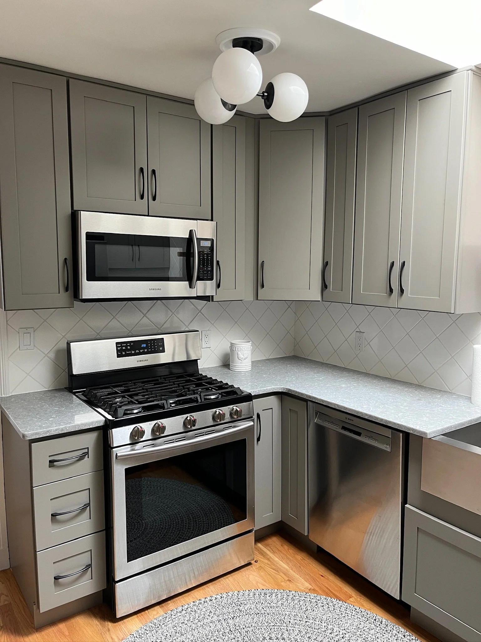 Kitchen with gray cabinets, stainless steel microwave above stove, stainless steel oven, built-in dishwasher, countertop, and a round woven rug on a wooden floor.