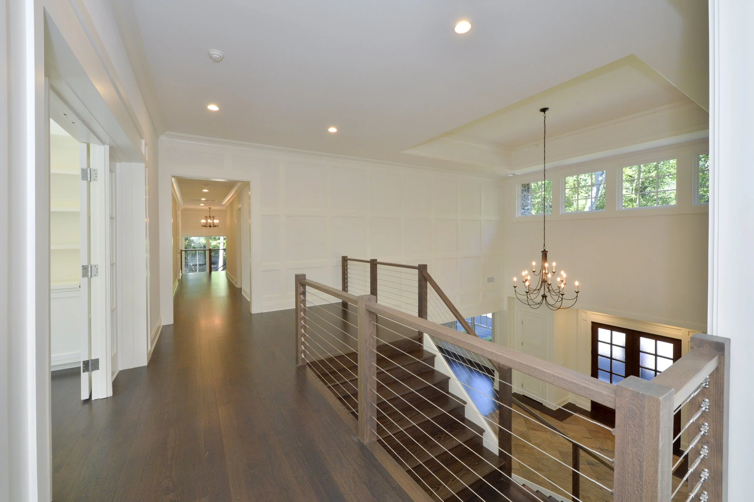 Interior view of a modern house with a wooden staircase, chandelier, white walls, large windows, hardwood floors, and hallway leading to other rooms.