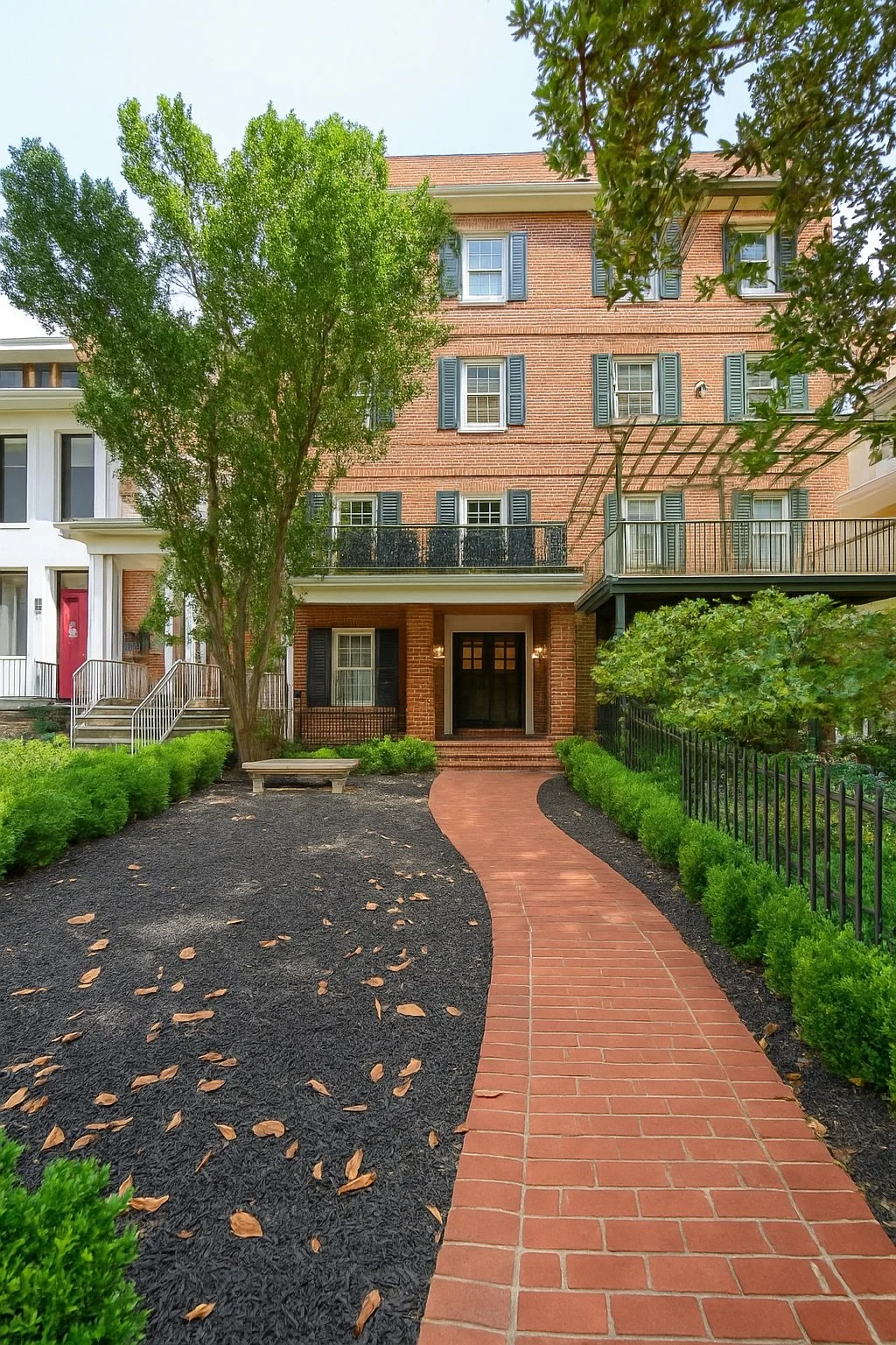 Red brick apartment building with black shutters, a small balcony, and arched front door, surrounded by greenery, a curved brick walkway, and a small landscaped garden.