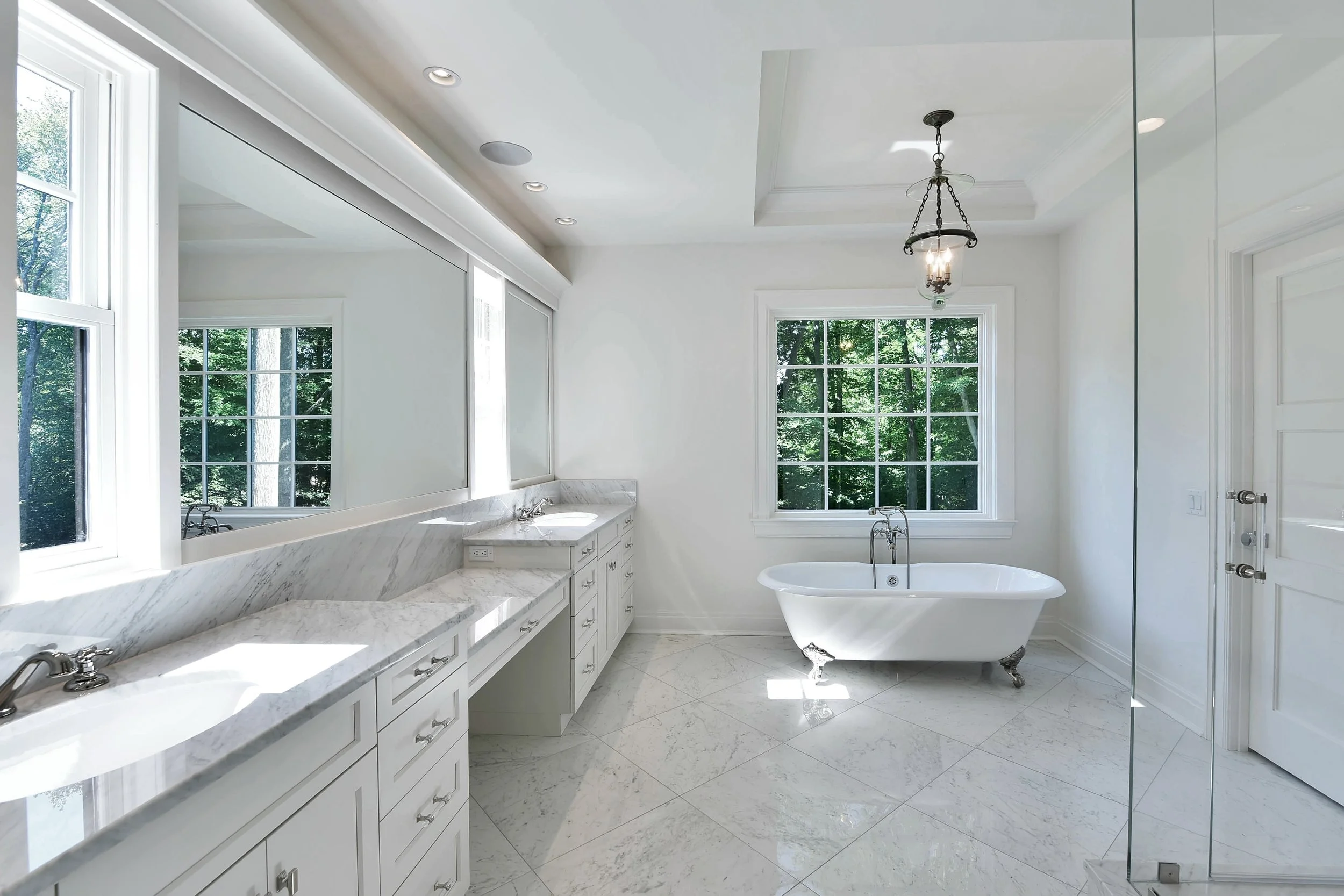 Bright bathroom with white marble countertops, a freestanding bathtub near large windows, and a walk-in shower with a glass door.
