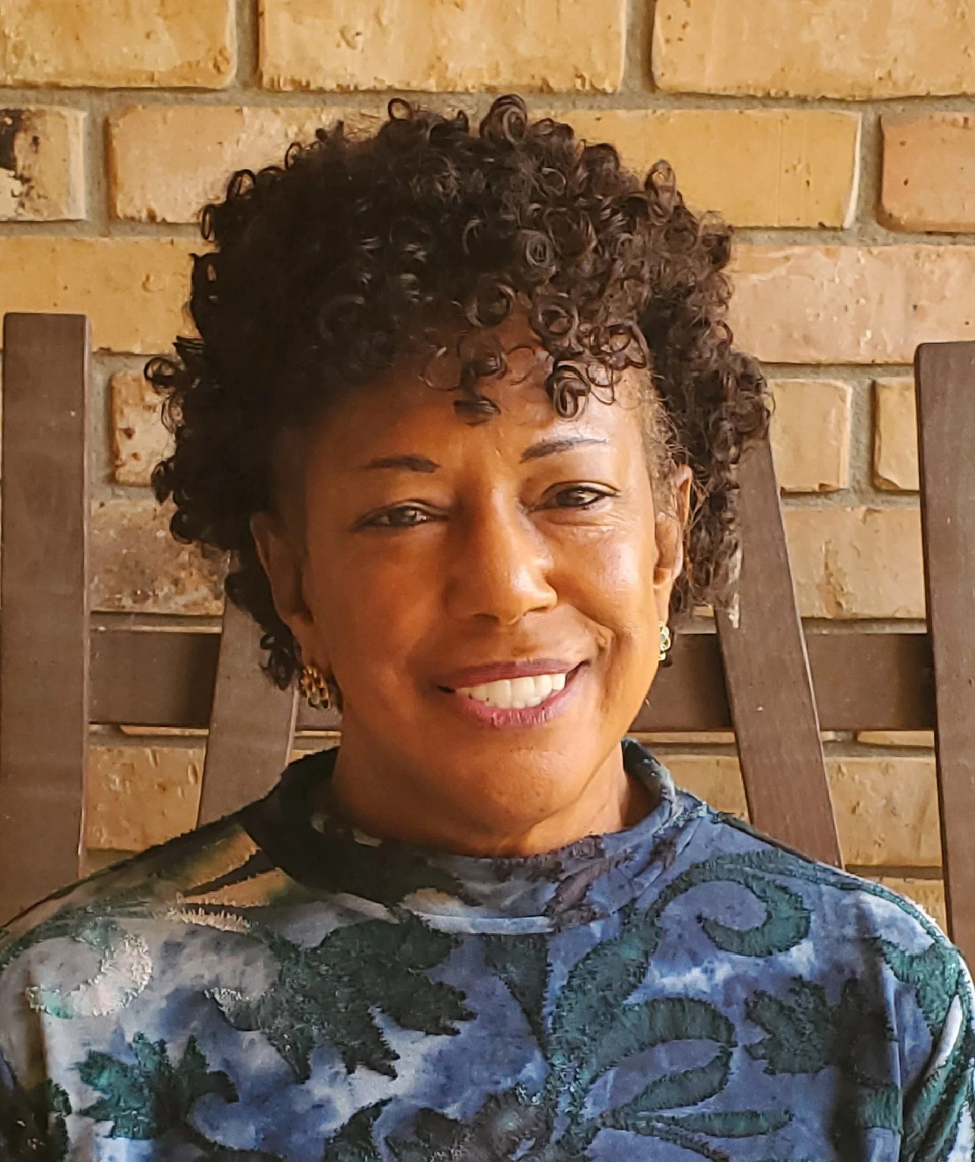 Dianne Mouton-Allen, NCBI Lafayette Chapter Director is smiling with curly hair, wearing a patterned top and earrings, sitting in front of a brick wall.