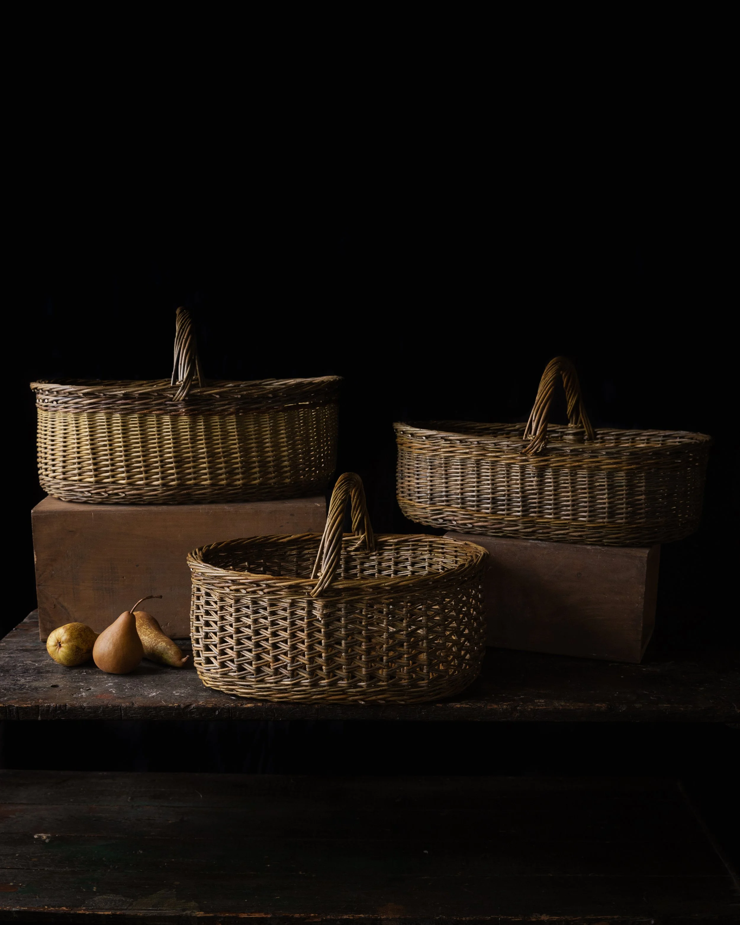 Three woven wicker baskets on a dark wooden surface, with three pears on the left side and a dark background.