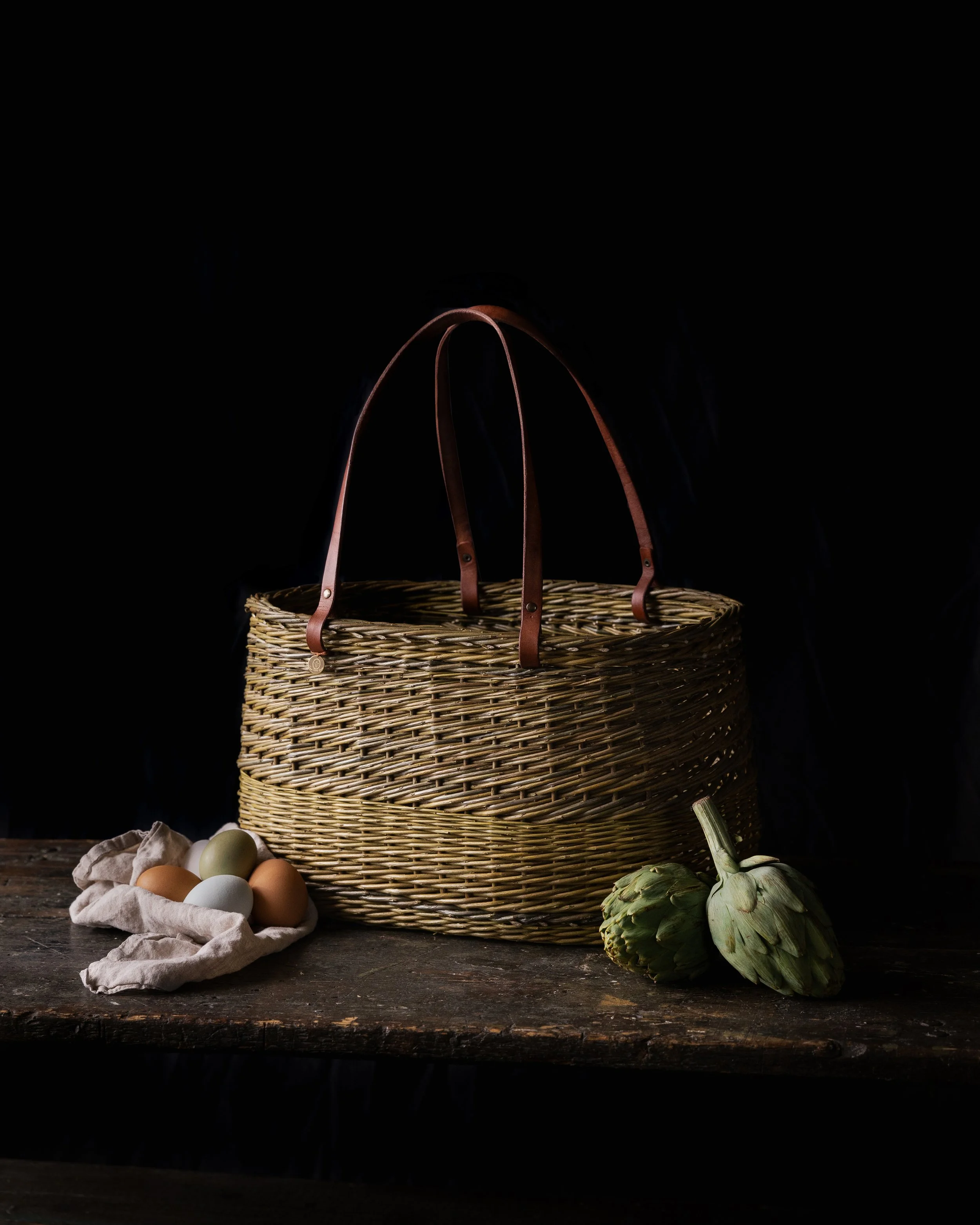 A wicker basket with leather handles, a cloth with eggs, and two artichokes on a rustic wooden table against a black background.