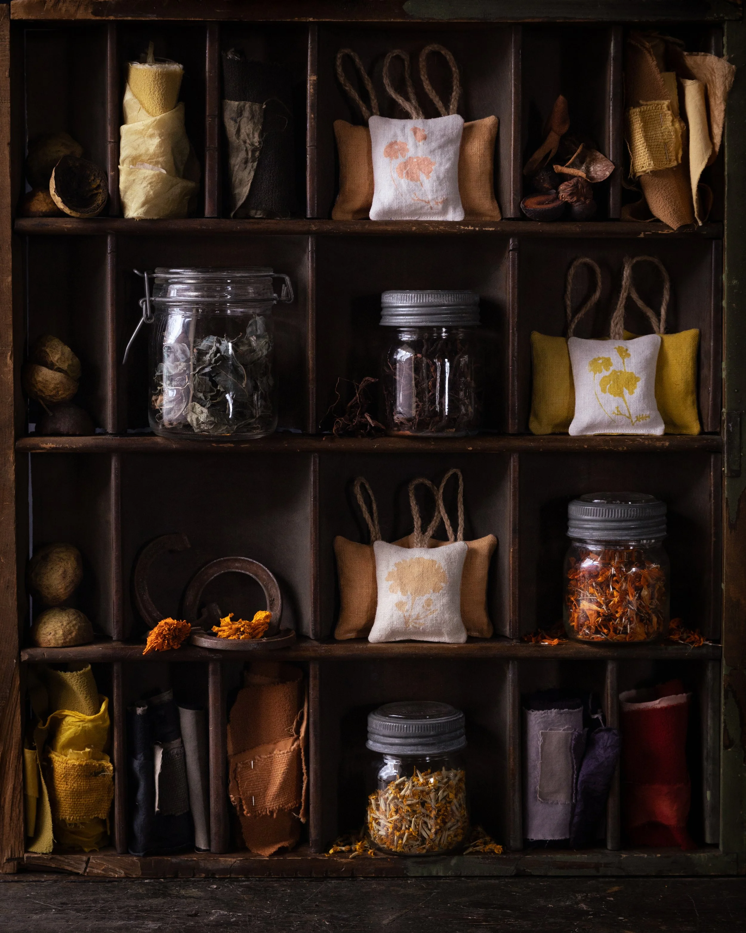 A vintage wooden cabinet with multiple compartments holding dried herbs, spices, and small embroidered linen pouches, some with floral designs.