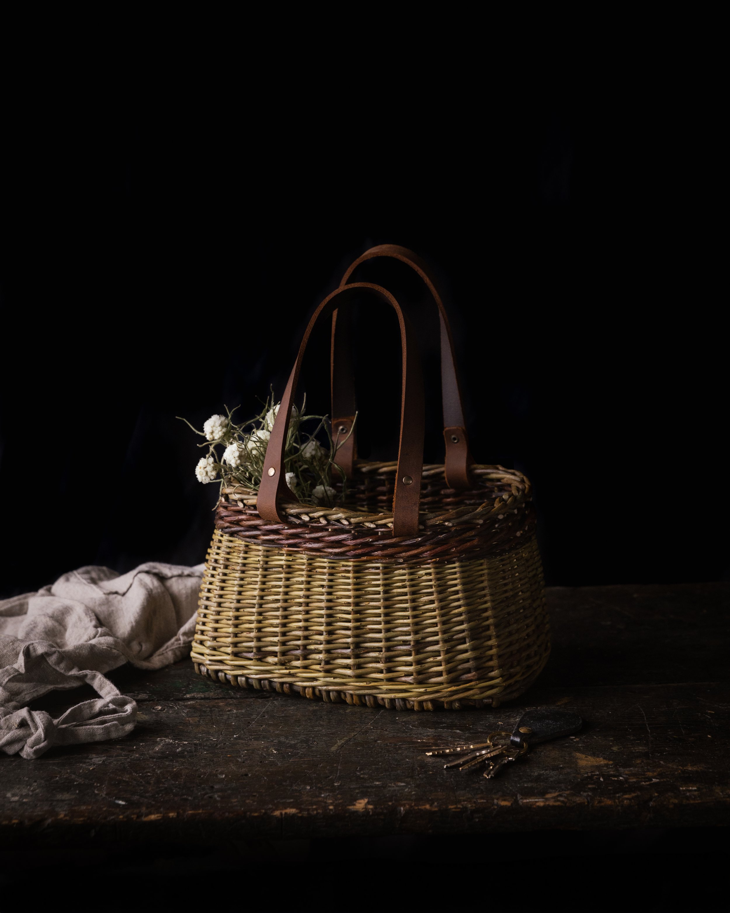 A woven basket with leather handles sitting on a rustic wooden table, with a bunch of small white flowers inside, and a set of keys beside it, against a dark background.