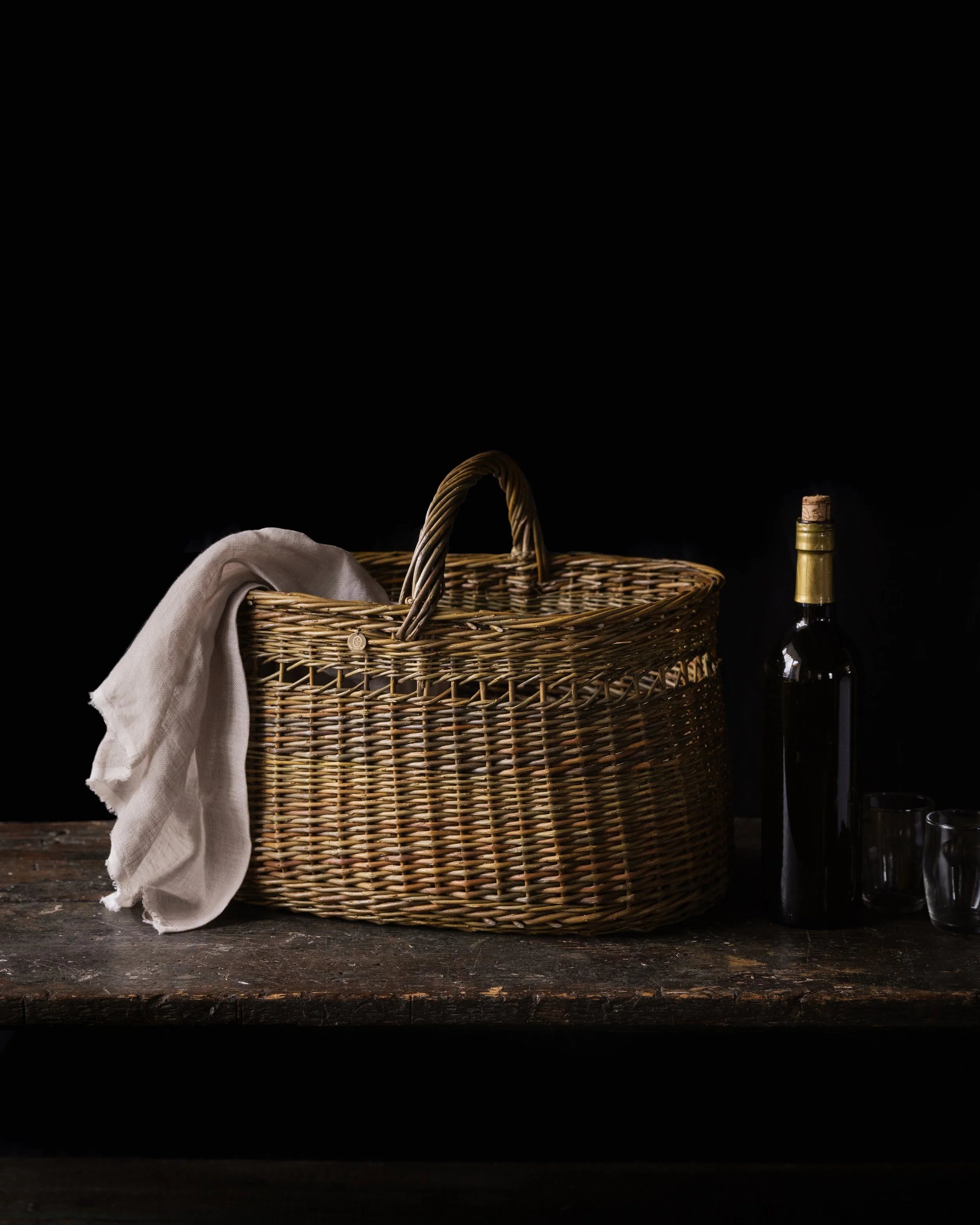 Wicker picnic basket with a white cloth draped over the side, a bottle of wine with a cork, and two empty glasses on a dark wooden surface against a black background.