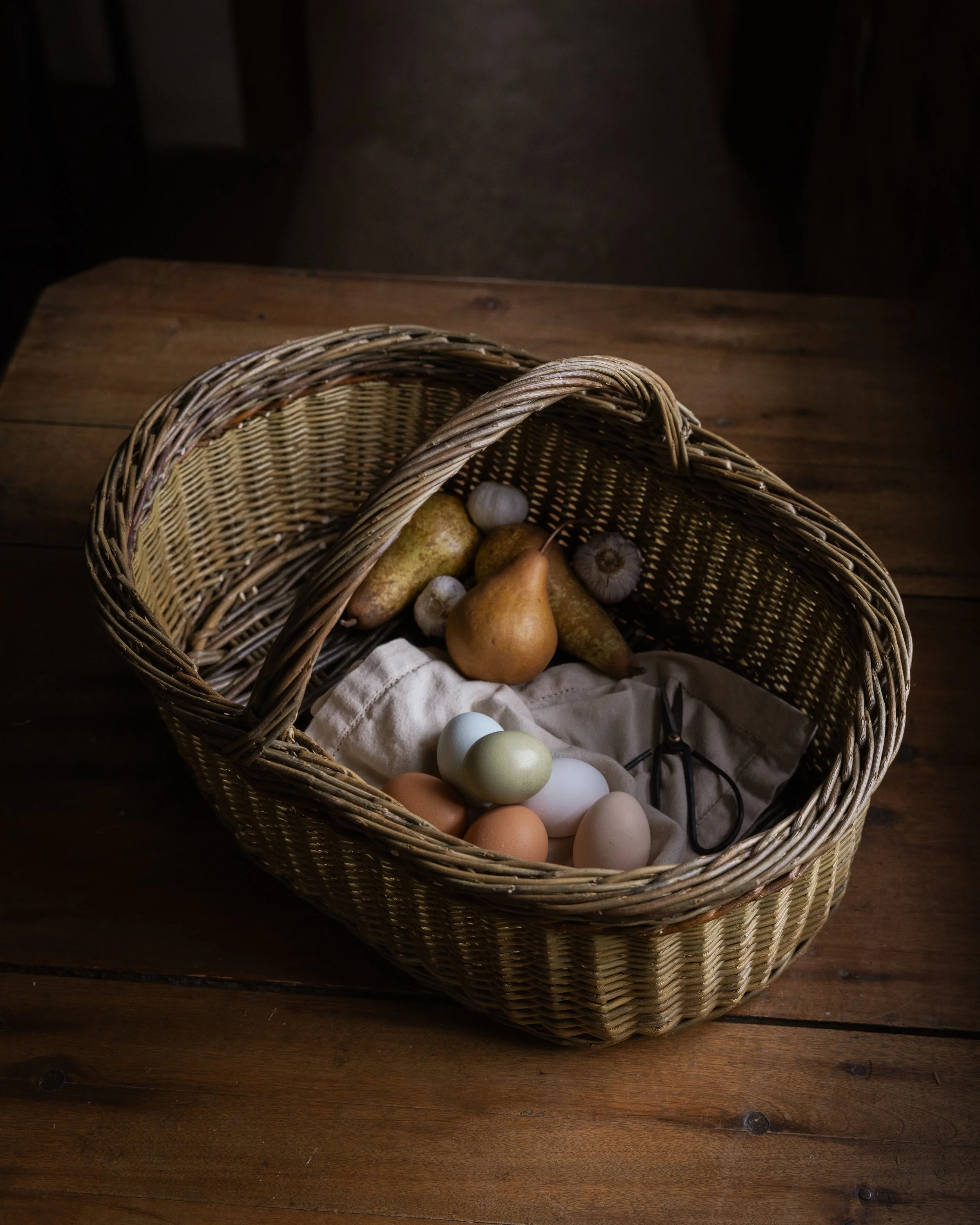 A woven wicker basket containing eggs, pears, garlic, and other vegetables on a wooden table.
