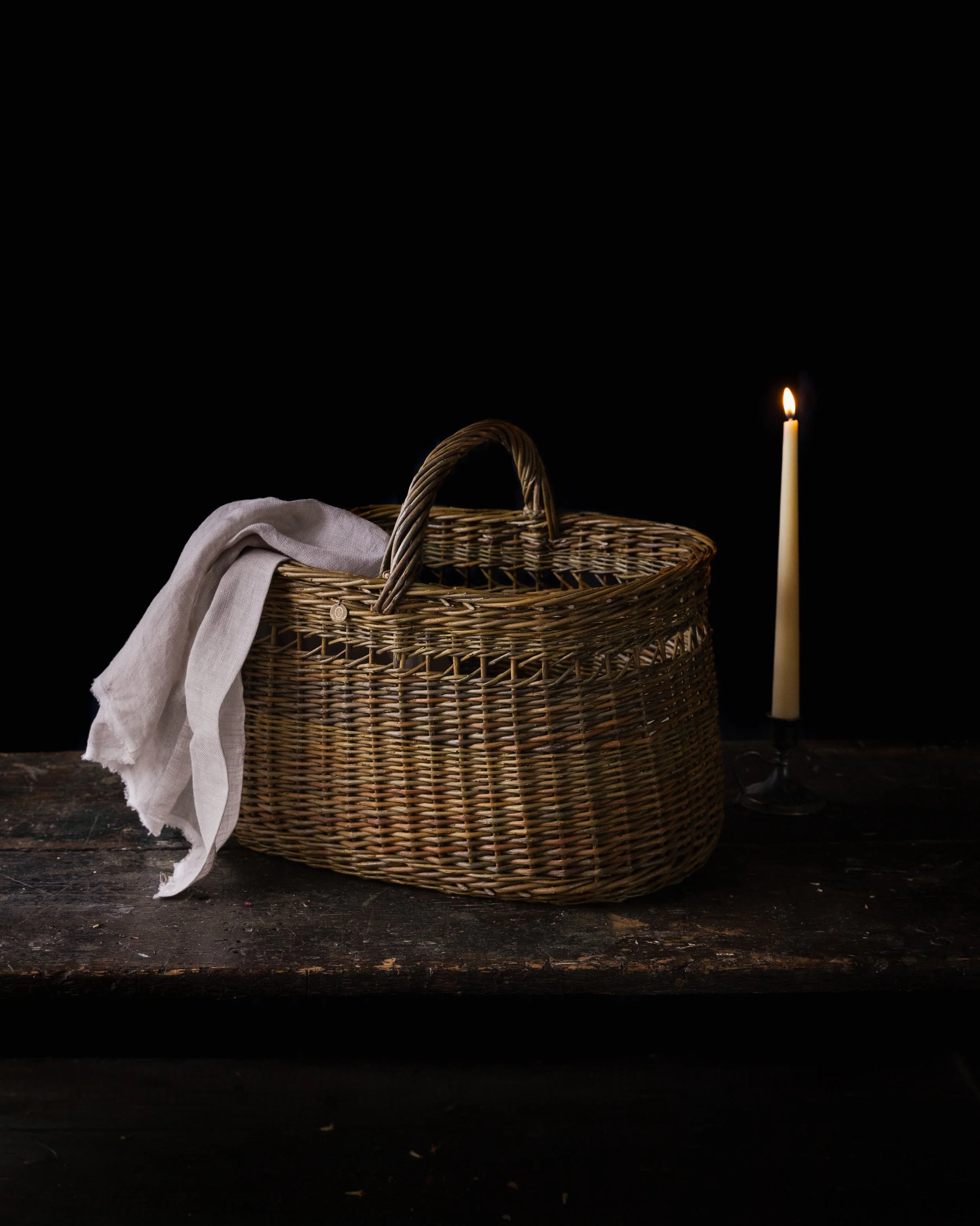 A wicker basket with a light-colored cloth draped over the edge, placed on a dark wooden surface, with a lit candle beside it and a black background.