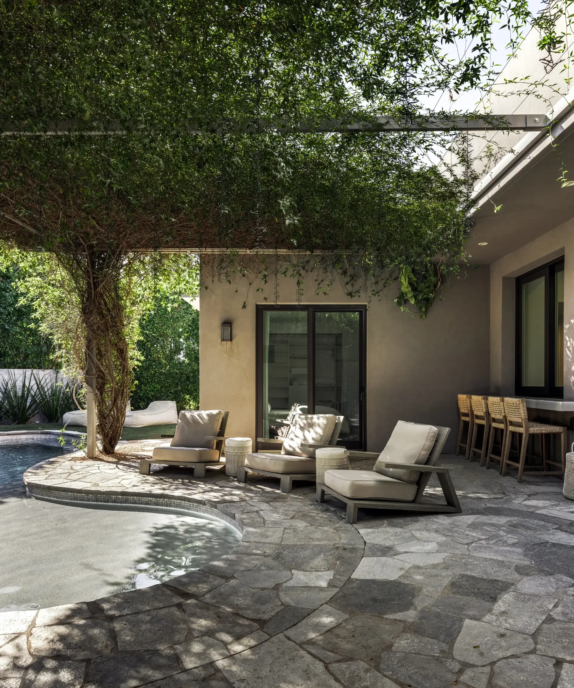 An outdoor patio with lounge chairs, side tables, a pool, and lush greenery overhead and around the area.