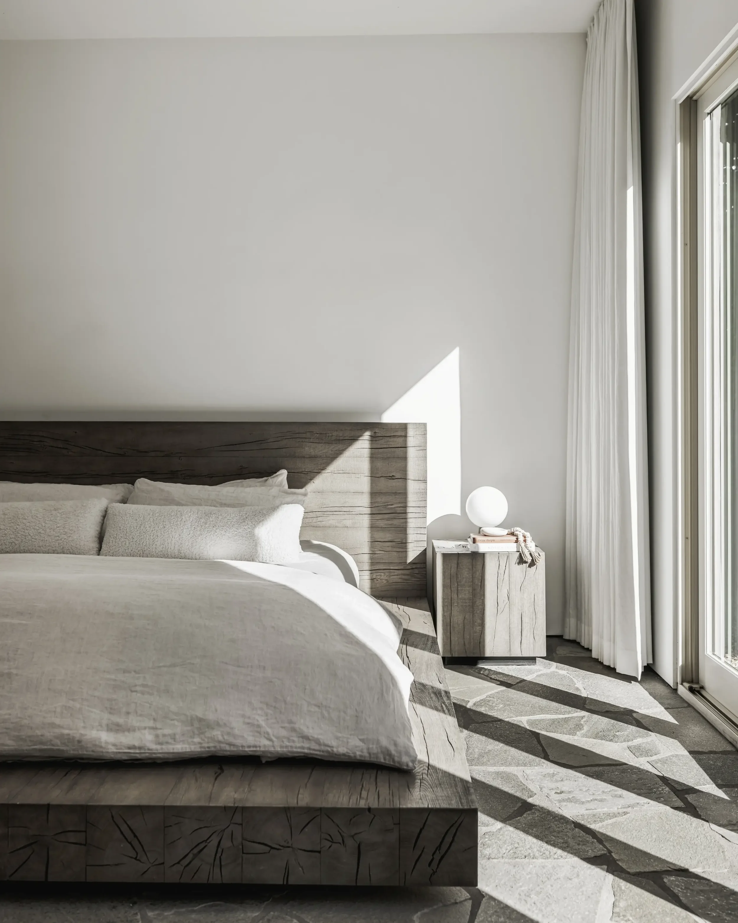 A minimalist bedroom with a wooden bed frame, white bedding, a nightstand with a round lamp, books, and decorative items, and a sliding glass door with white curtains allowing sunlight to cast shadows on the floor.