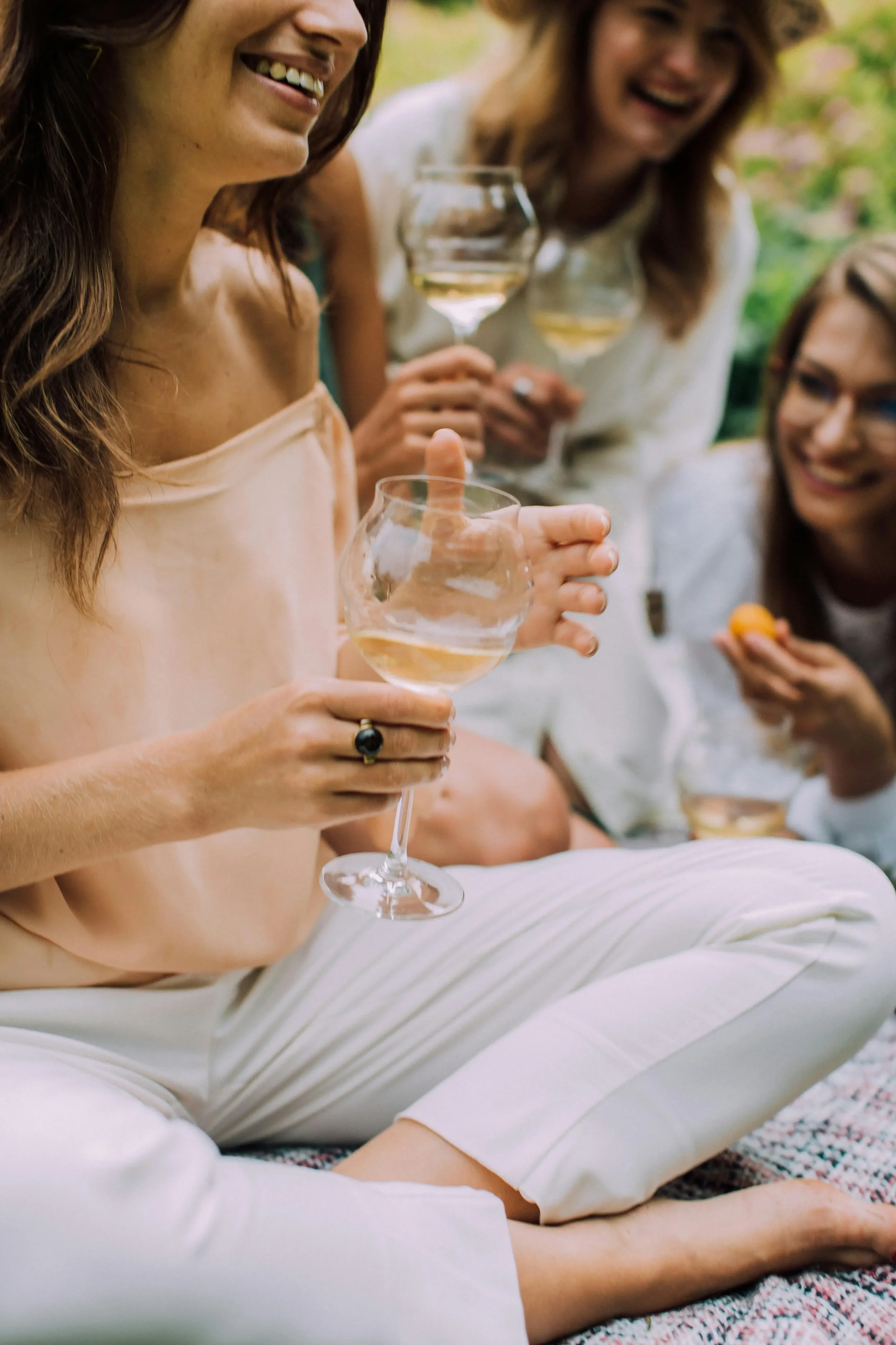 A woman in a peach-colored top and white pants sitting outdoors on a blanket, holding a glass of white wine, surrounded by friends who are also holding wine glasses, smiling and enjoying a picnic in a lush green setting.