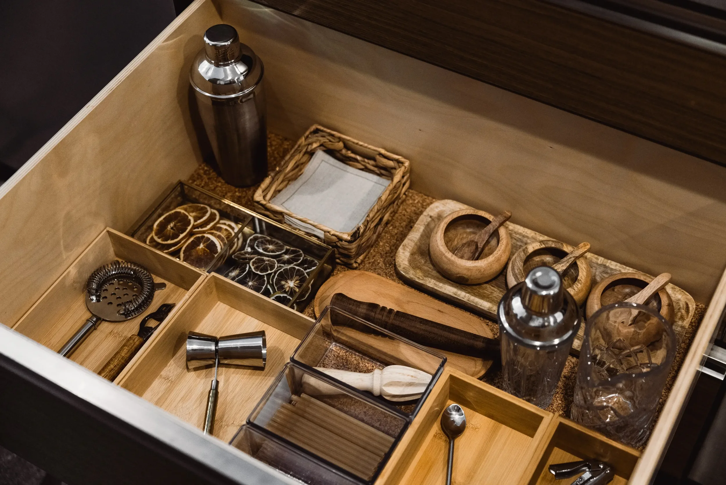 Open drawer with bar tools, dried citrus slices, small bowls with spoons, and glassware.