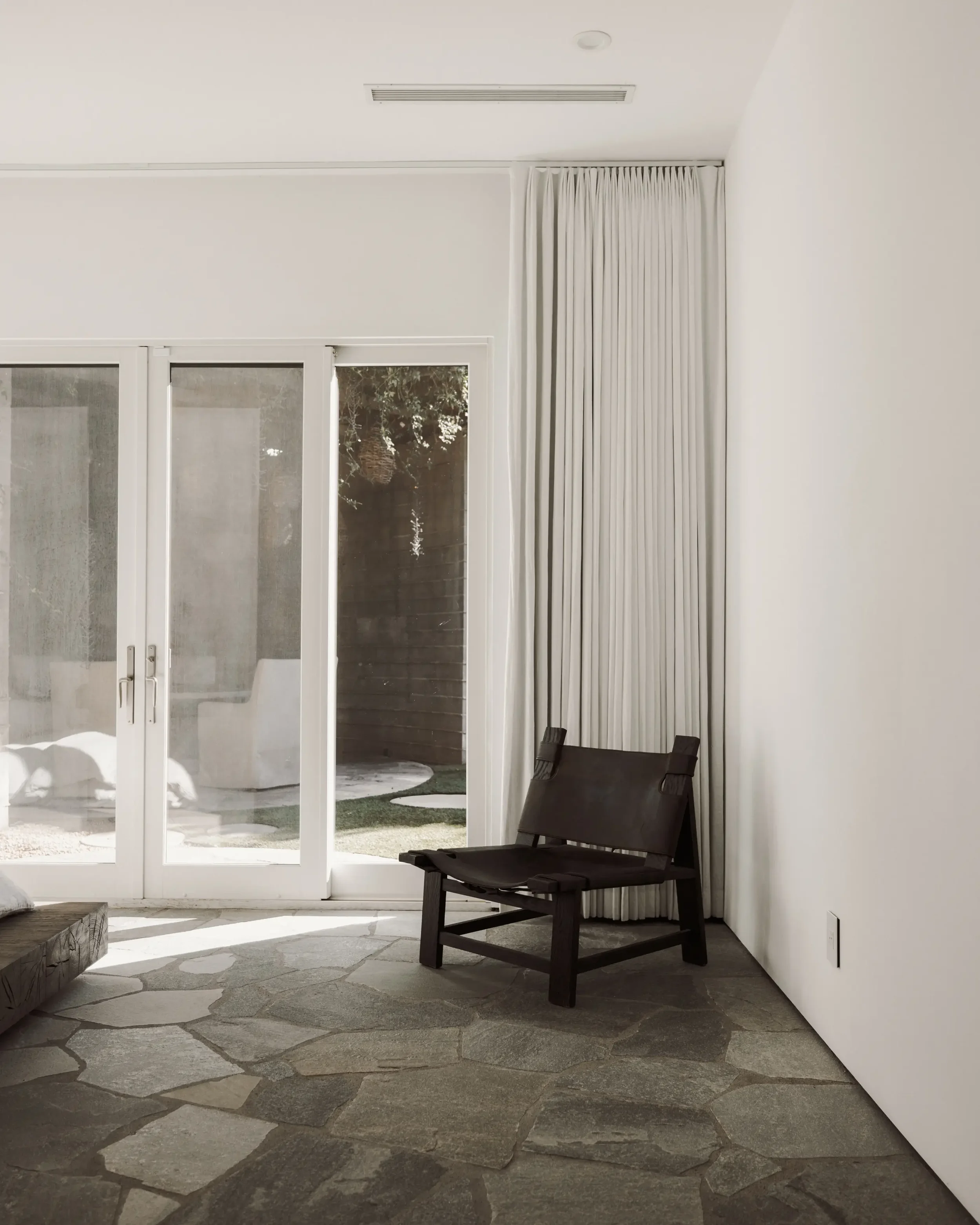 A corner of a modern interior room with a black wooden chair and a stone floor, near glass sliding doors and white curtains, with a view of the outdoor patio with a dog.