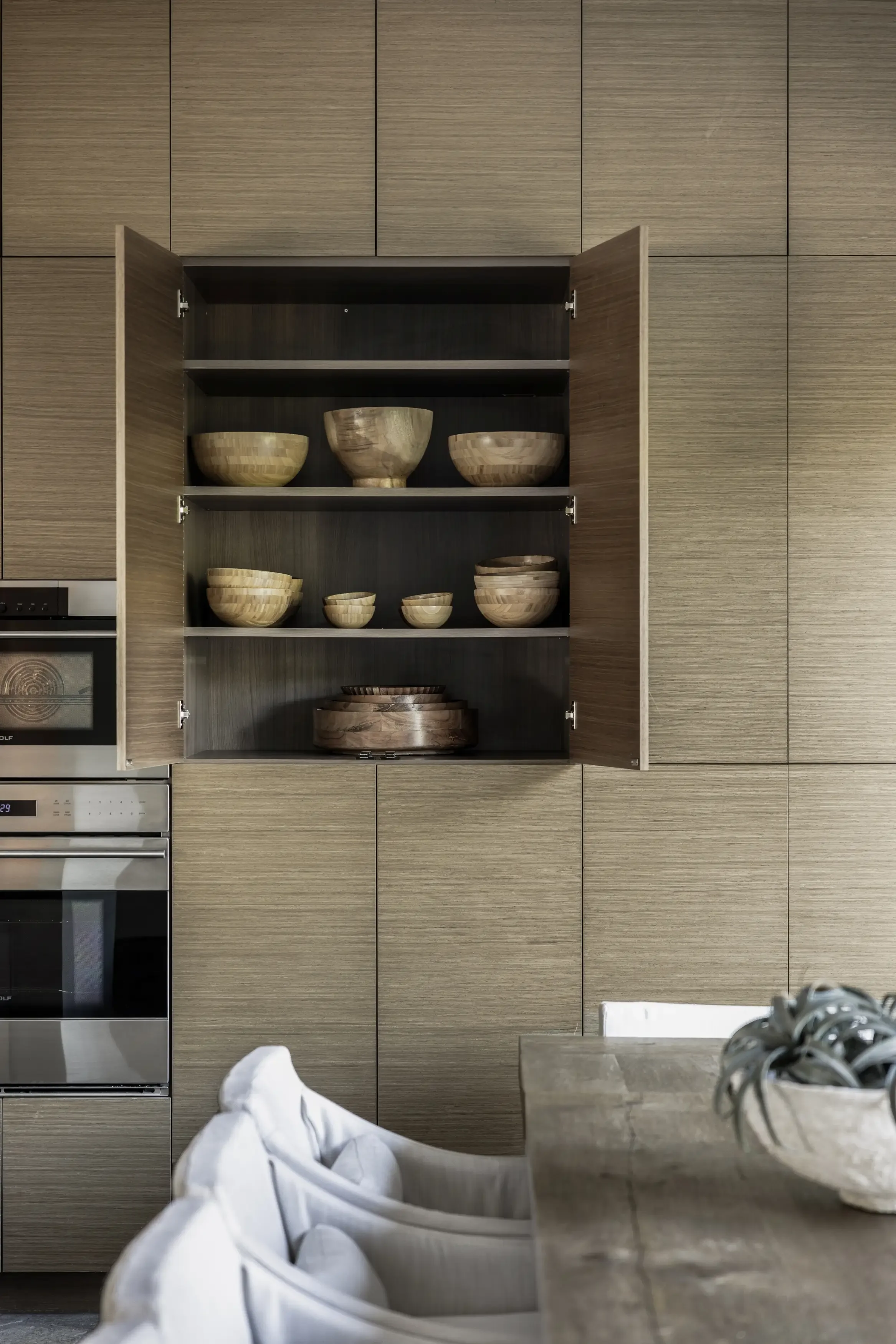 Open kitchen cabinet with wooden bowls, set against a wood-paneled wall, with part of a dining table with white chairs and a decorative bowl in the foreground.