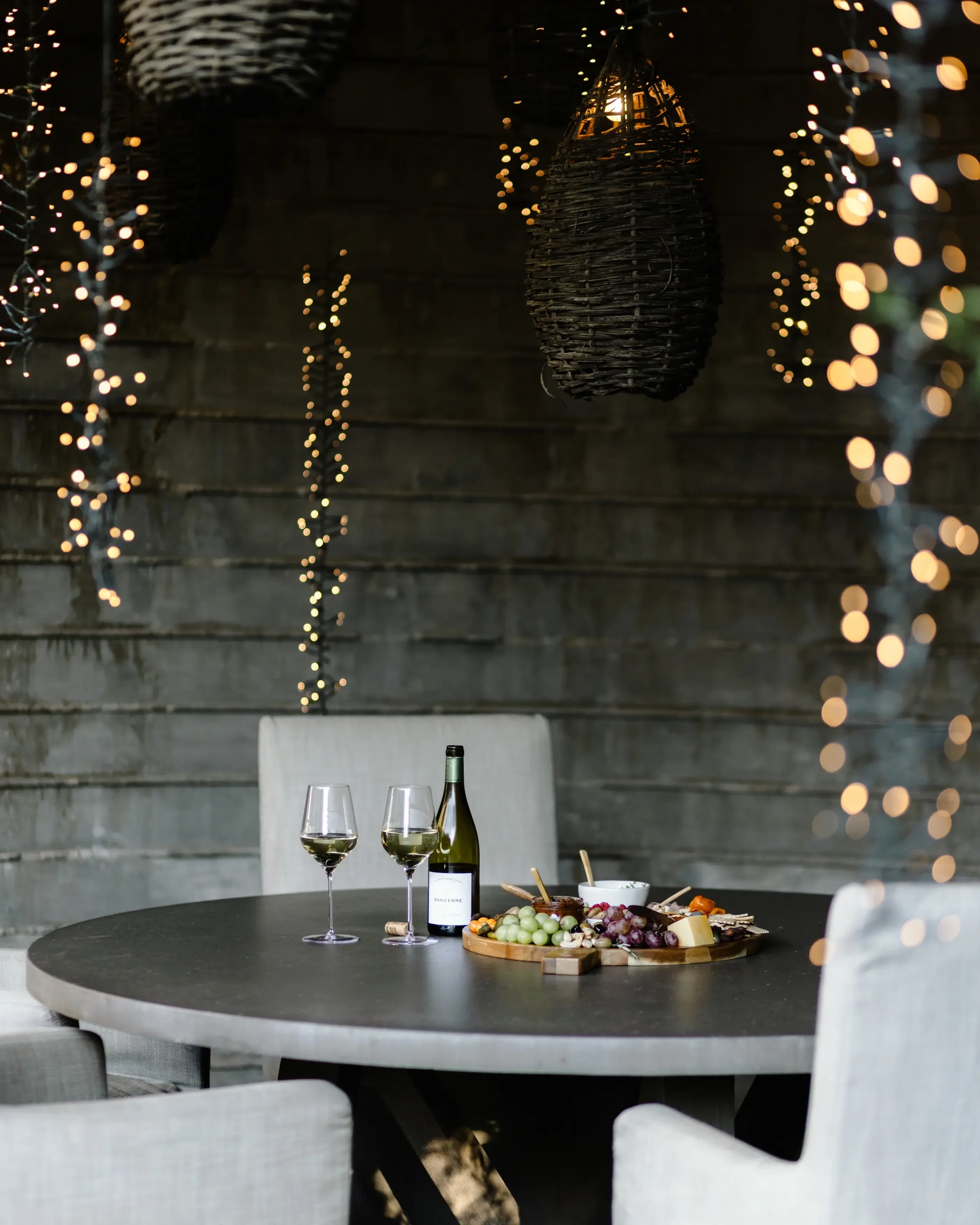 A dining table with two glasses of white wine, a bottle of white wine, and a charcuterie board with grapes and assorted cheeses in a cozy, dimly lit room with string lights and woven pendant lamps.