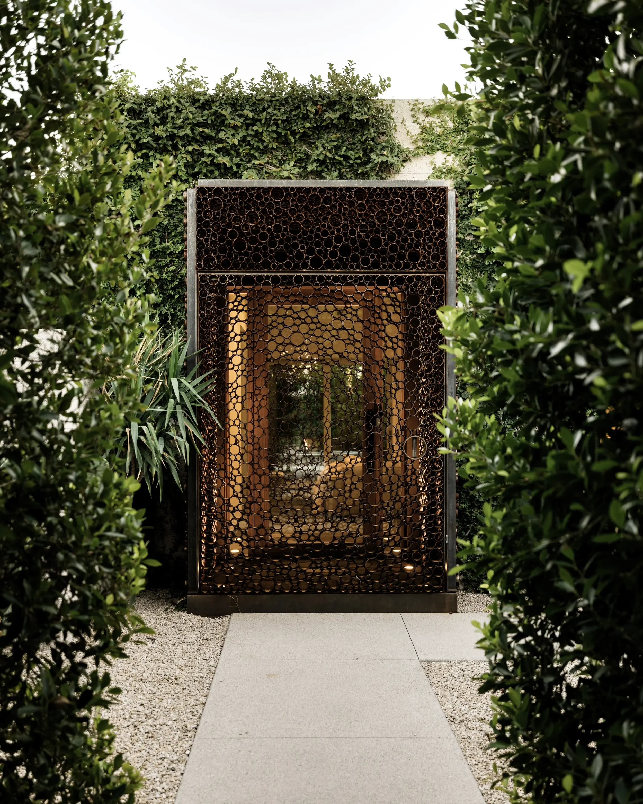 Modern iron gate with circular cut-out design, framed by green shrubbery, leading to a courtyard with outdoor furniture.