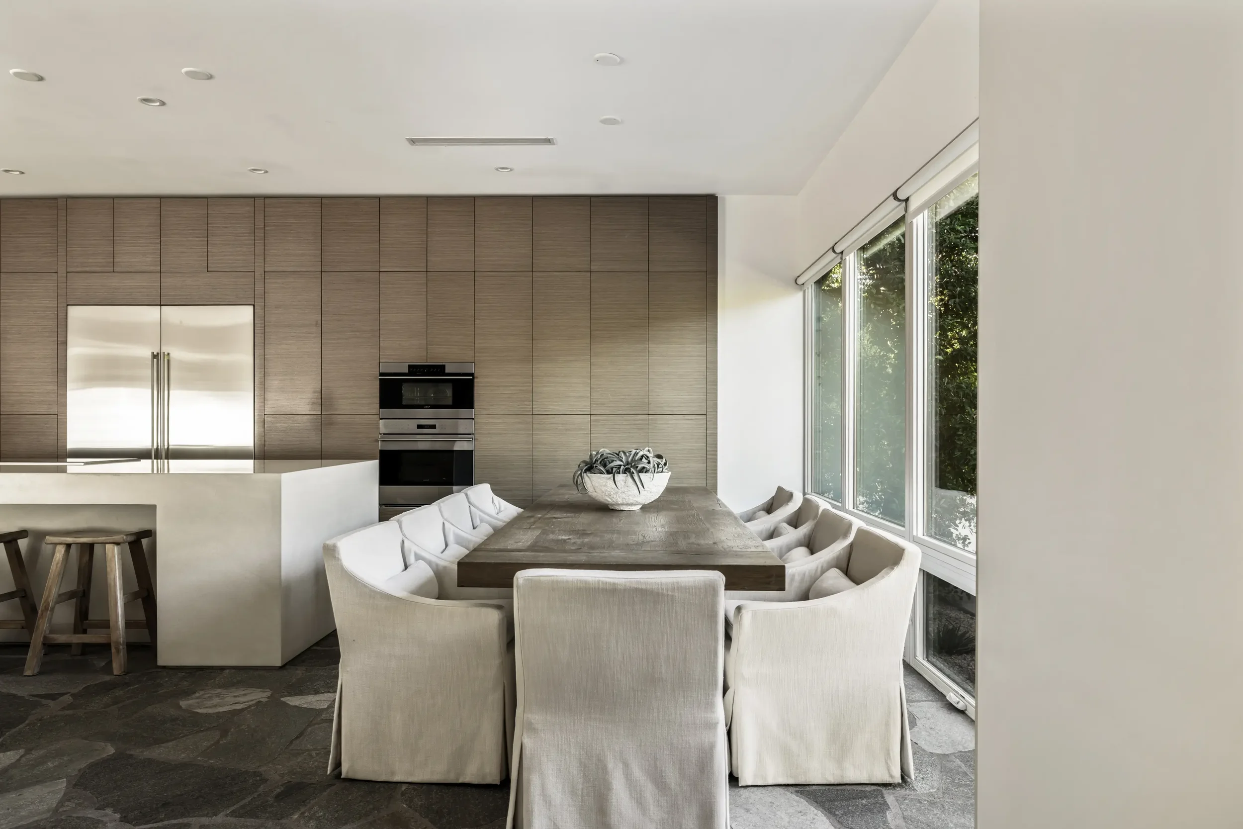 Modern dining room with large wooden table surrounded by cream-colored upholstered chairs, large windows with greenery outside, and a built-in wooden wall with stainless steel appliances.