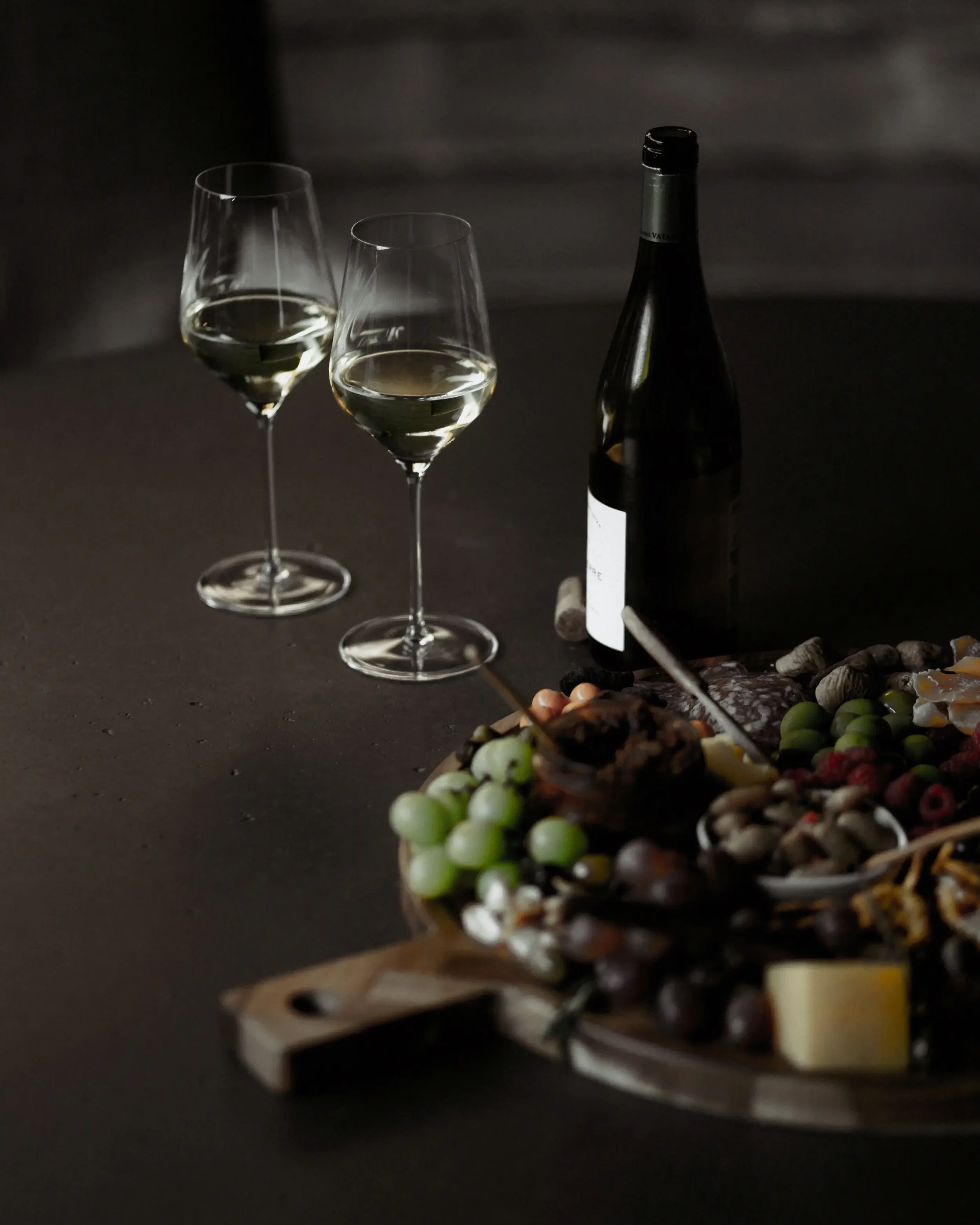 Two glasses of white wine, a bottle of wine, and a charcuterie board with grapes, cheese, and snacks on a dark table.