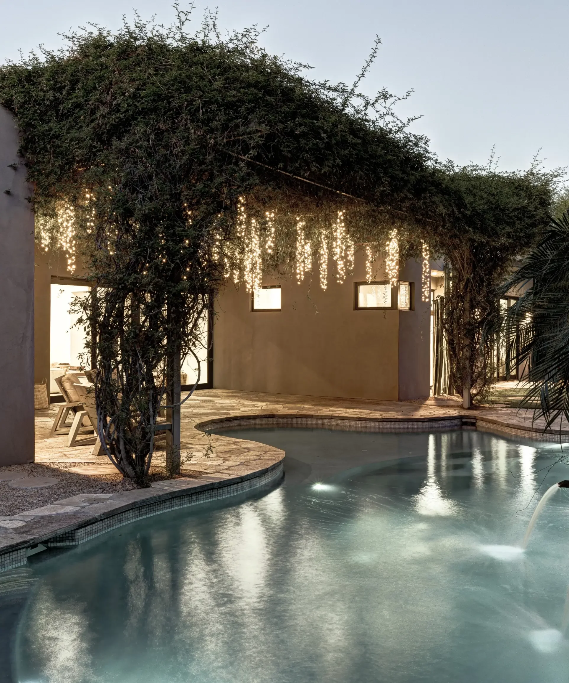 A backyard pool area at dusk with string lights hanging from a vine-covered pergola above the pool.