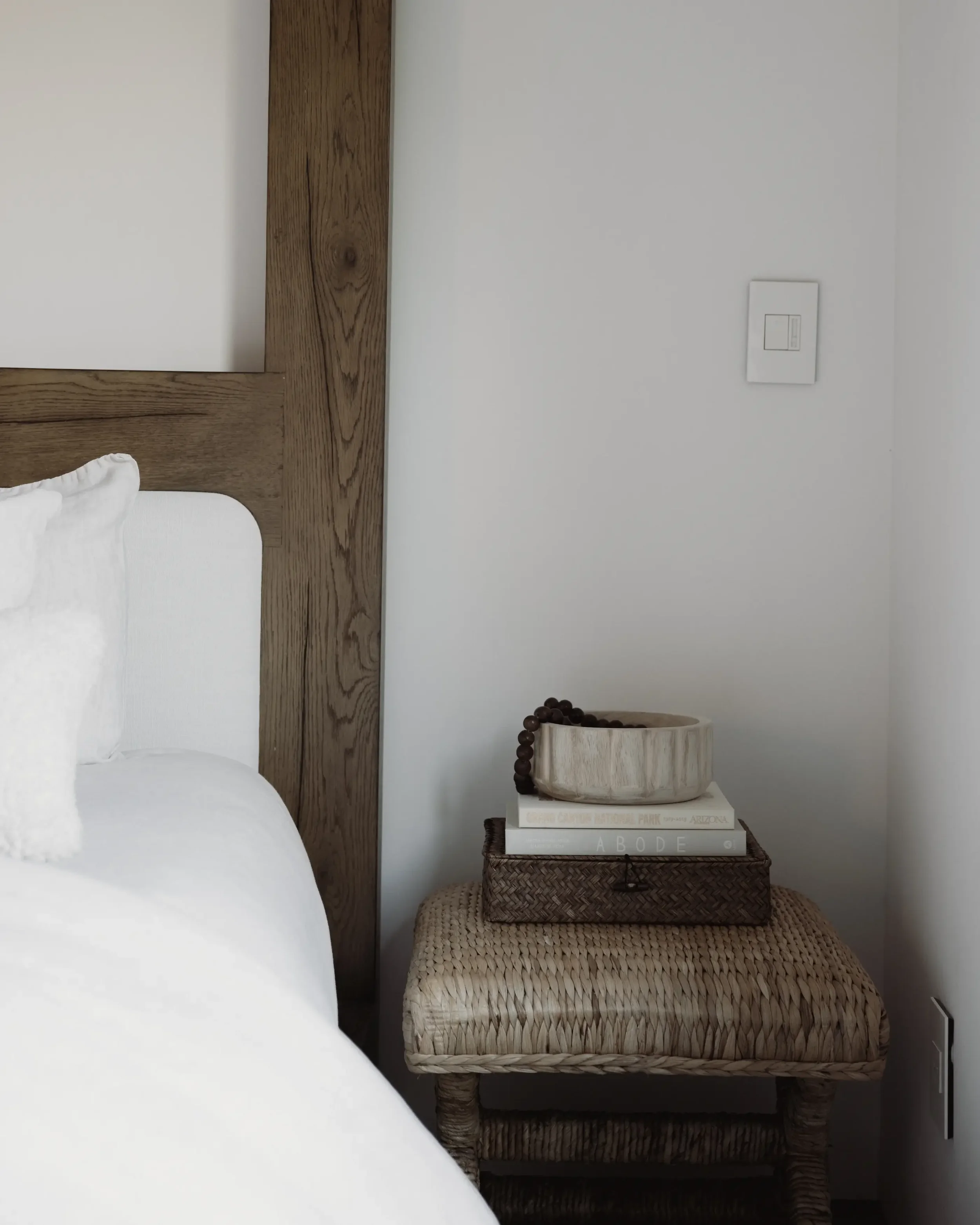 Bedroom corner with a white bed, wooden headboard, woven side table holding books and decorative objects.