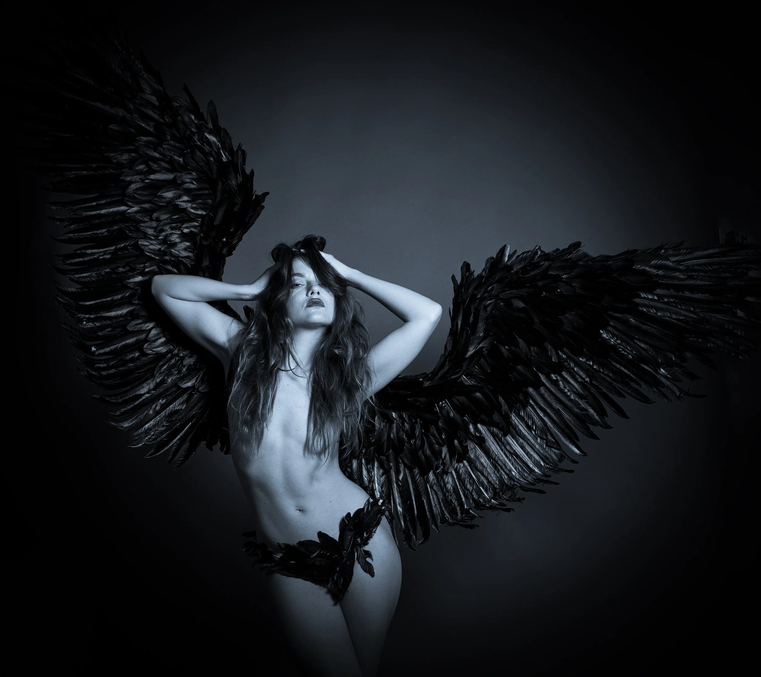 A woman with long hair and dark lipstick poses topless with her arms behind her head, wearing large black feathered wings.