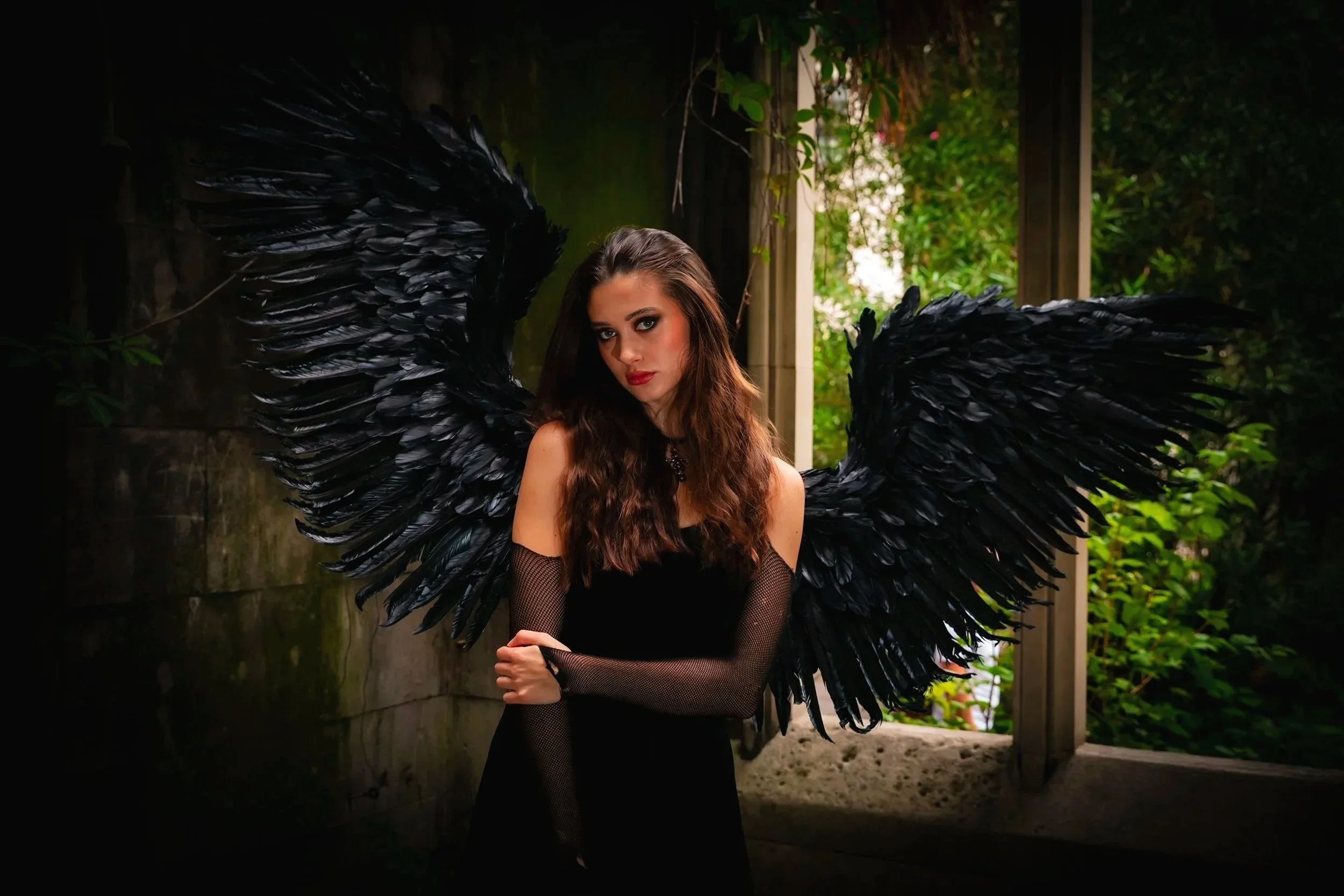 A young woman with long wavy brown hair, wearing a black dress and fishnet gloves, standing outdoors with black angel wings on her back.