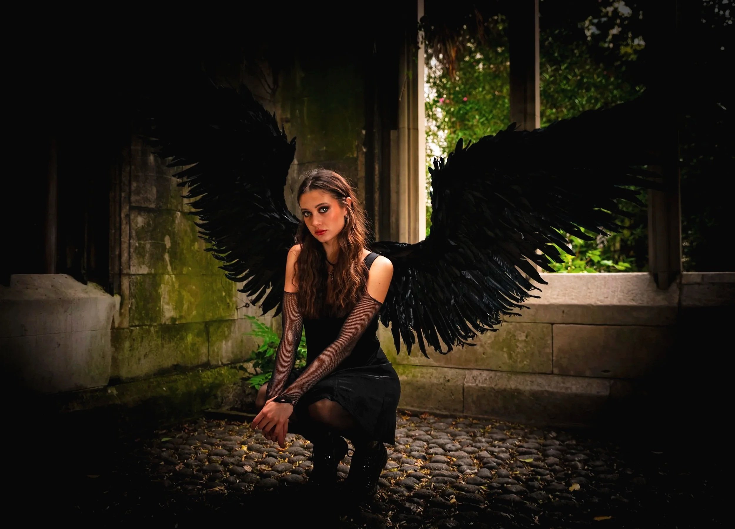 A woman with long wavy brown hair dressed in black, wearing black mesh gloves, with large black angel wings, kneeling on cobblestone ground in a dimly lit, outdoor stone alcove with greenery outside.