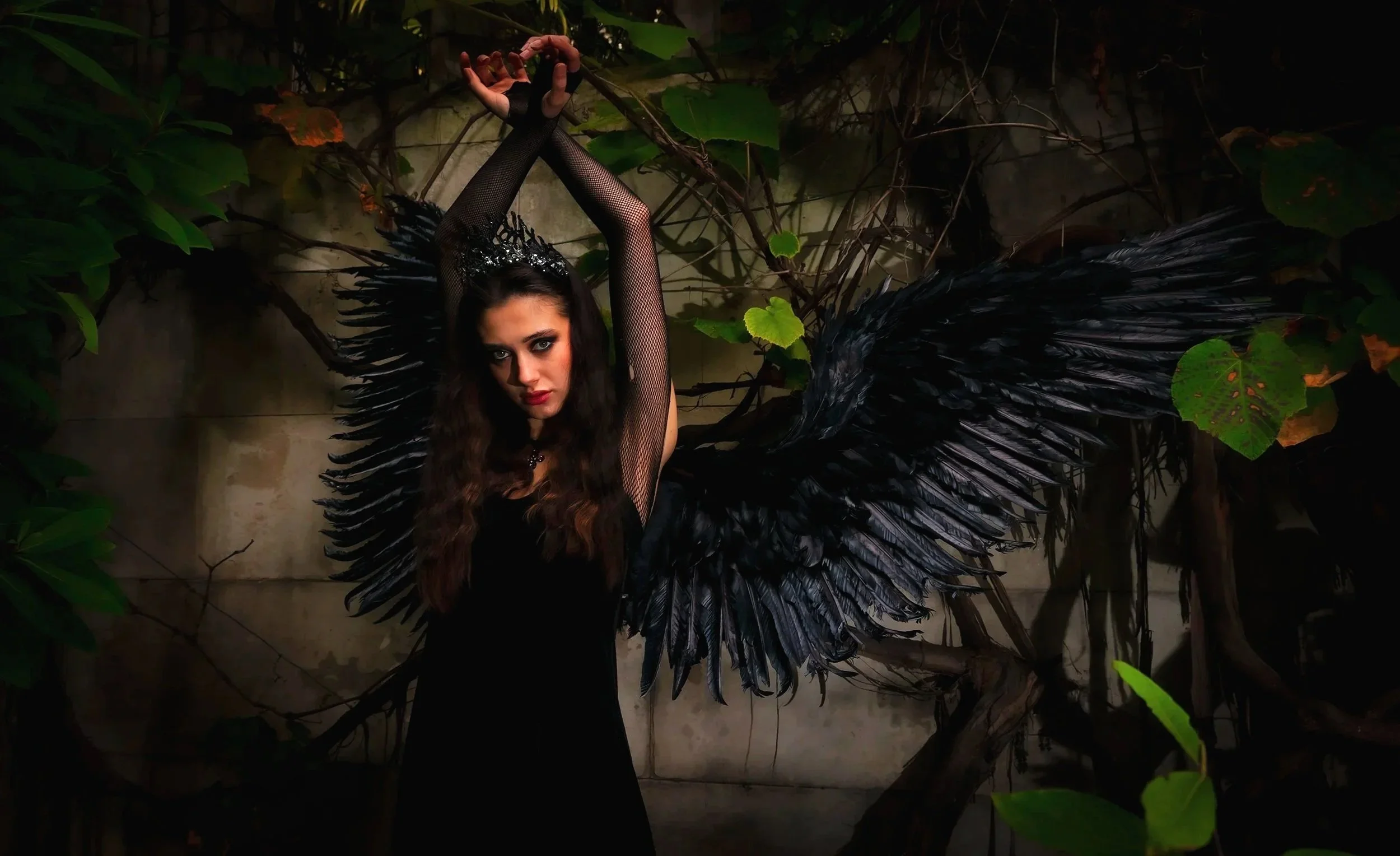 A woman dressed as a dark angel or demon with large black wings, wearing a black dress, fishnet sleeves, and a crown, standing in front of a wall with vines and leaves.