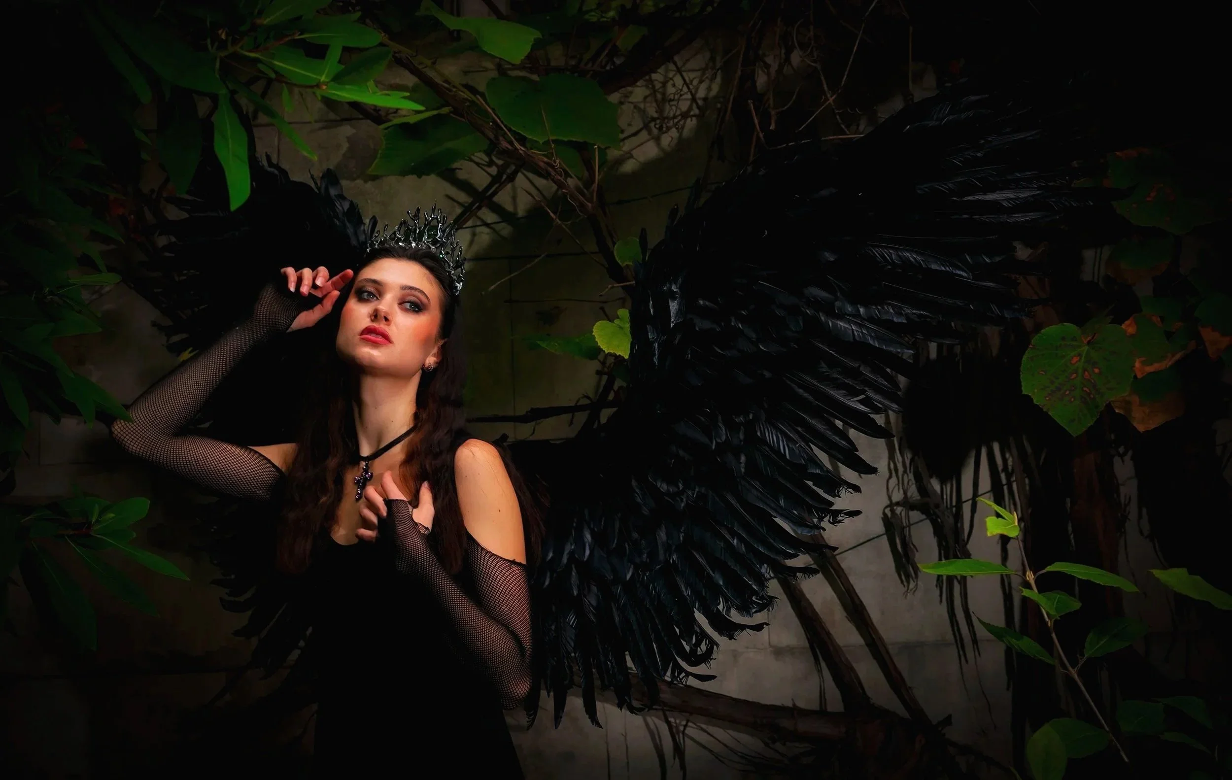 A woman dressed as a dark angel with black wings, a crown, and gothic accessories, posing amidst green foliage in a dimly lit environment.