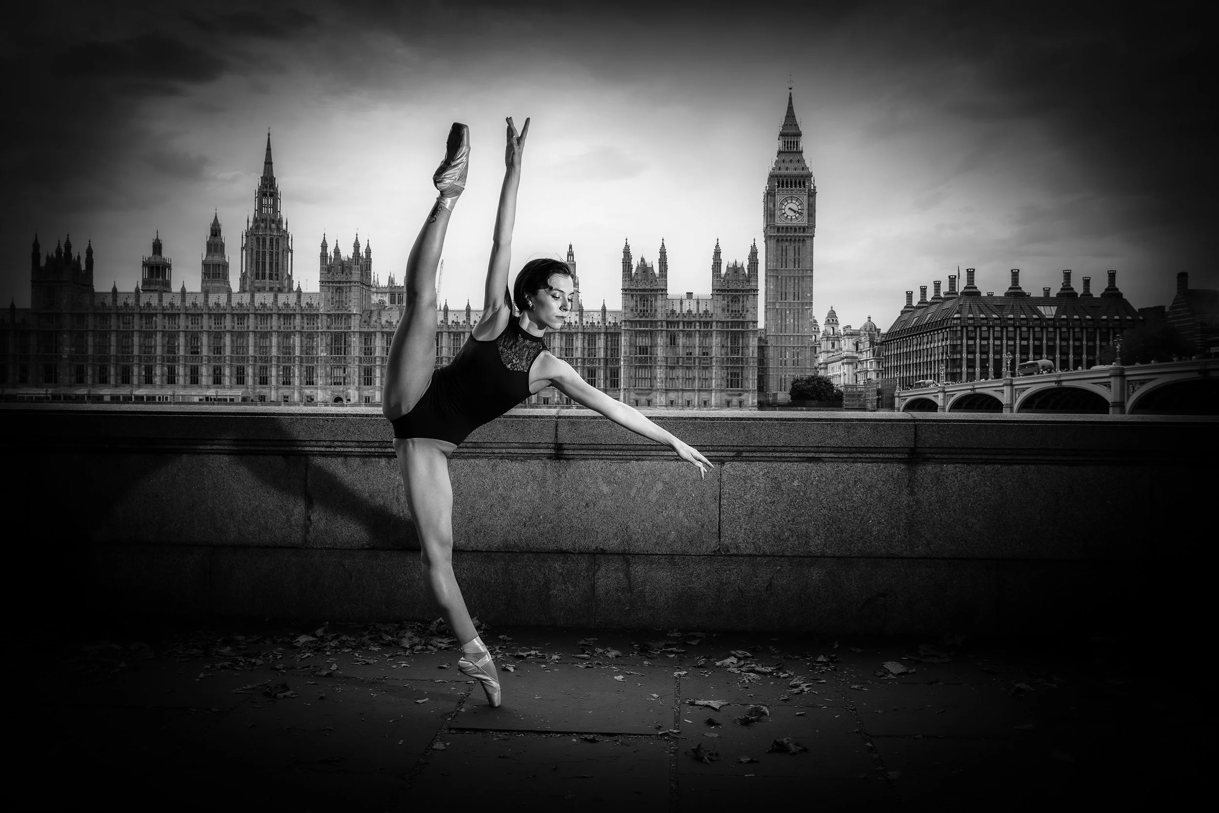 A ballet dancer performing a pose in front of a cityscape featuring the Big Ben clock tower and historic buildings in London, black and white photograph.
