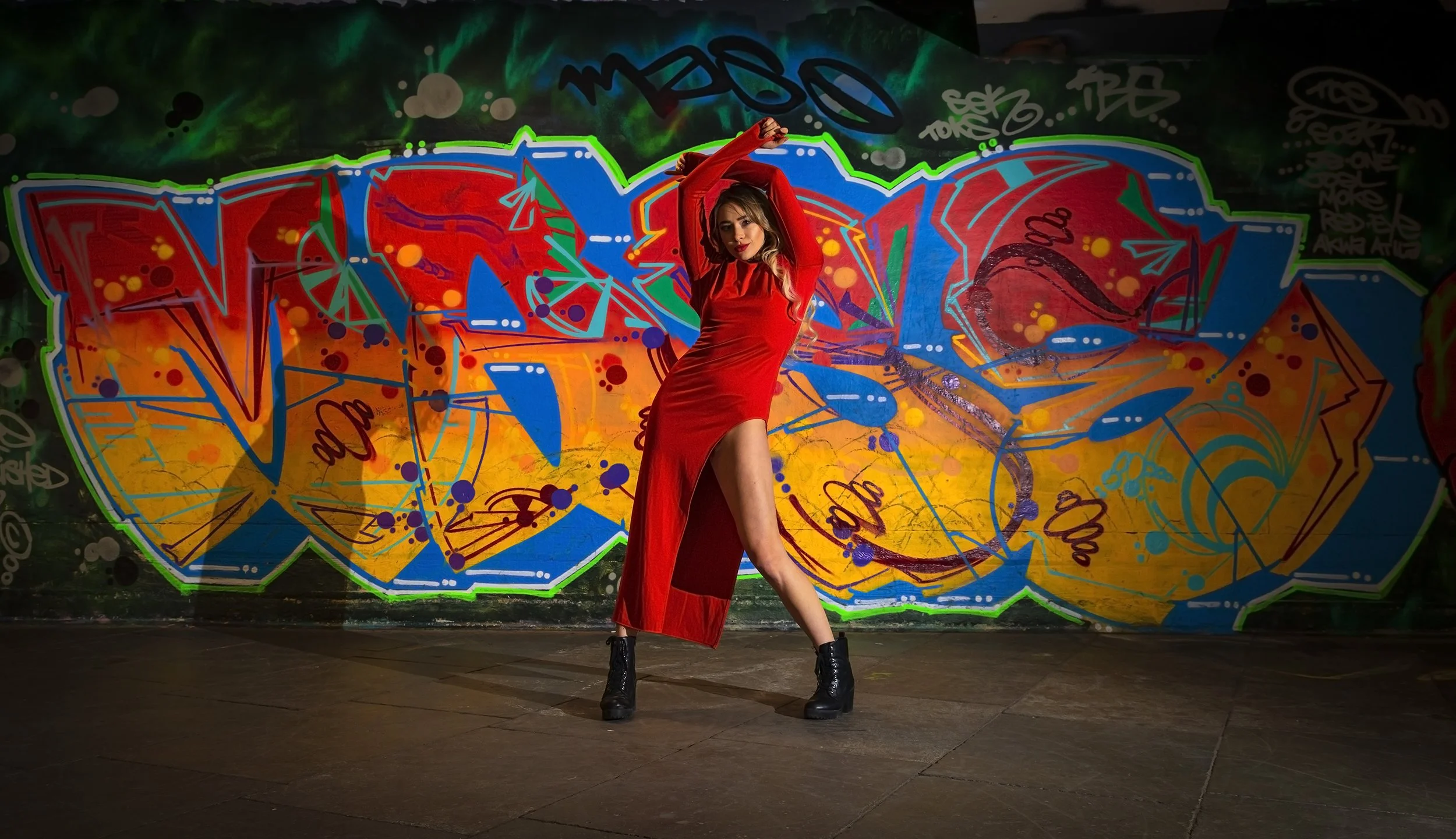 Young woman in a red dress and black boots posing in front of colorful graffiti wall.