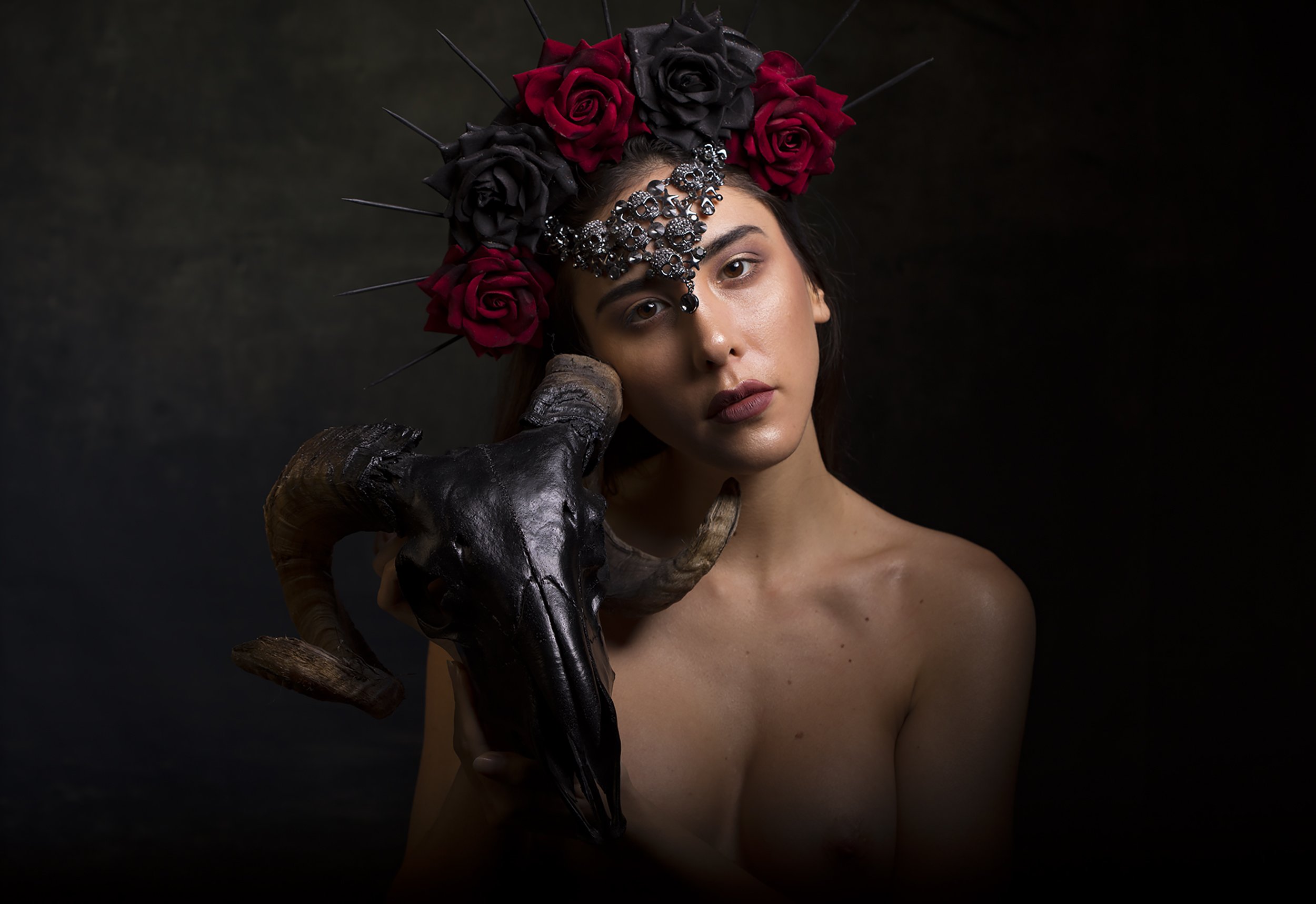 A woman with a dark floral crown and a decorative silver facial jewelry, posing topless with a black horned animal skull resting against her face, against a dark background.