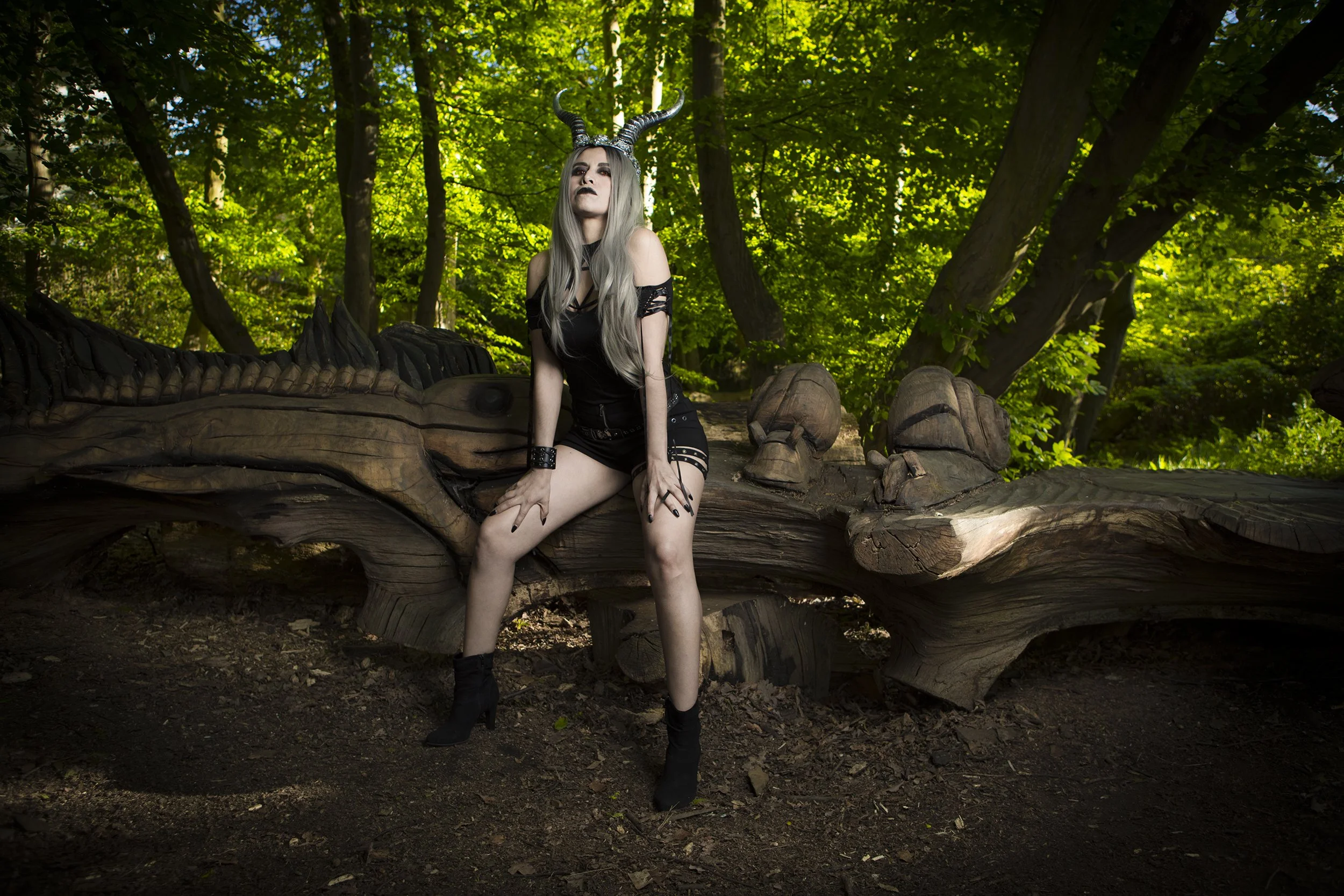 A person with silver hair, horns, dark makeup, and goth clothing sitting on a carved wooden sculpture in a forest with green foliage.