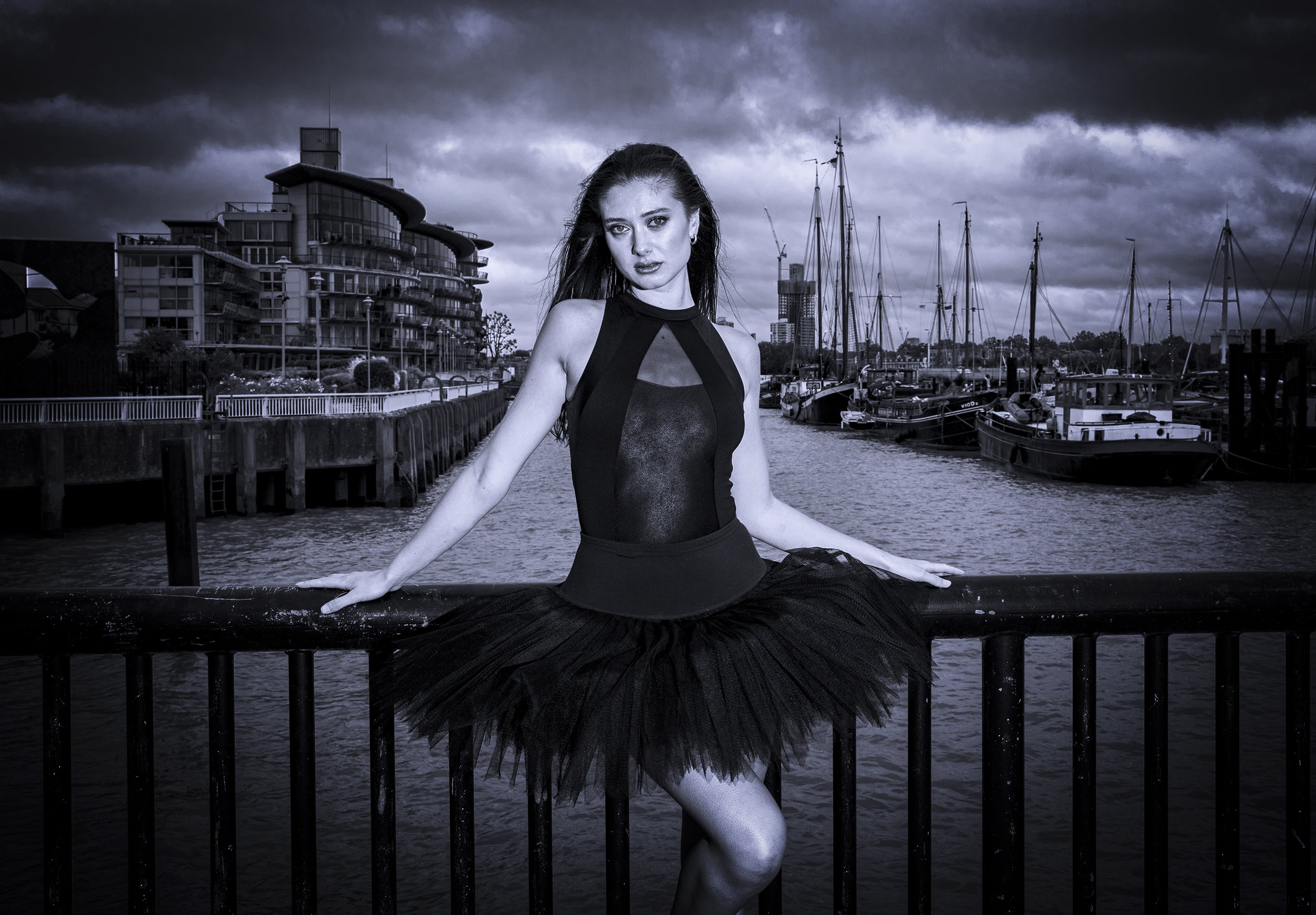 A woman in a black ballet tutu and sleeveless top sitting on a dock by the water with boats and modern buildings in the background, under a dark cloudy sky.