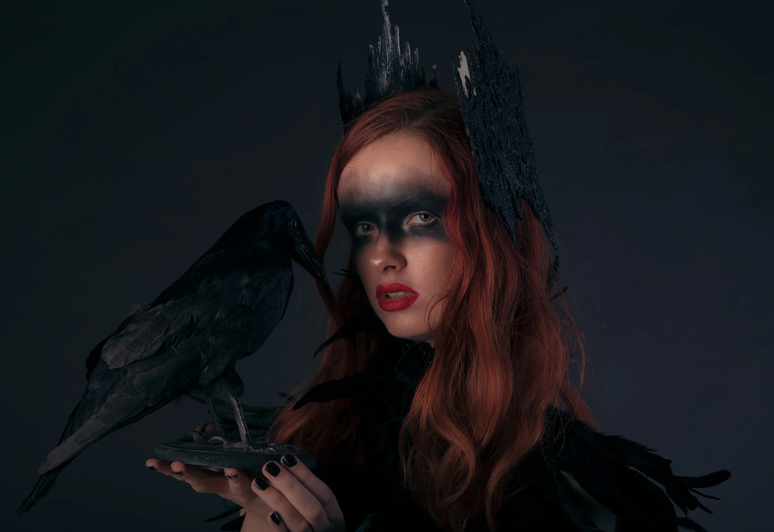A woman with long red hair dressed as a dark sorceress or witch, wearing a crown-like headpiece, with dark makeup, black dress, holding a black raven on her hand, against a dark background.