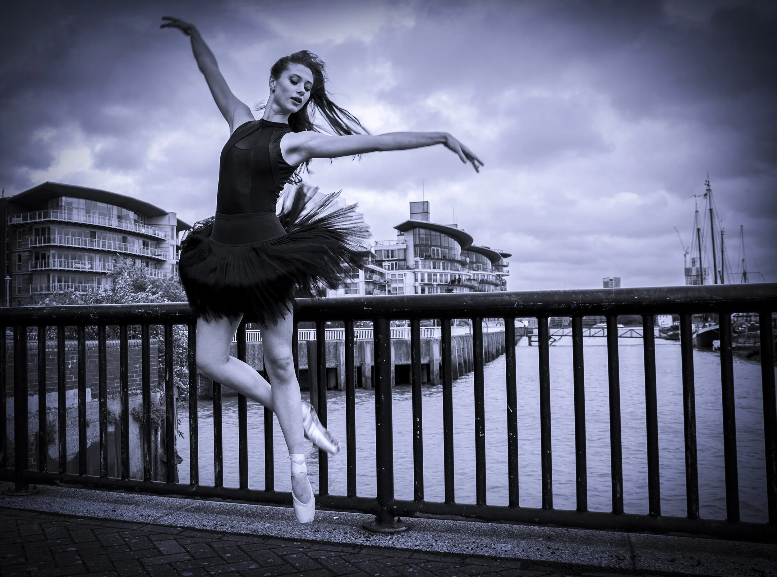 Ballet dancer posed on a waterfront with modern buildings and boats in the background, wearing a black tutu and pointe shoes, captured in black and white.