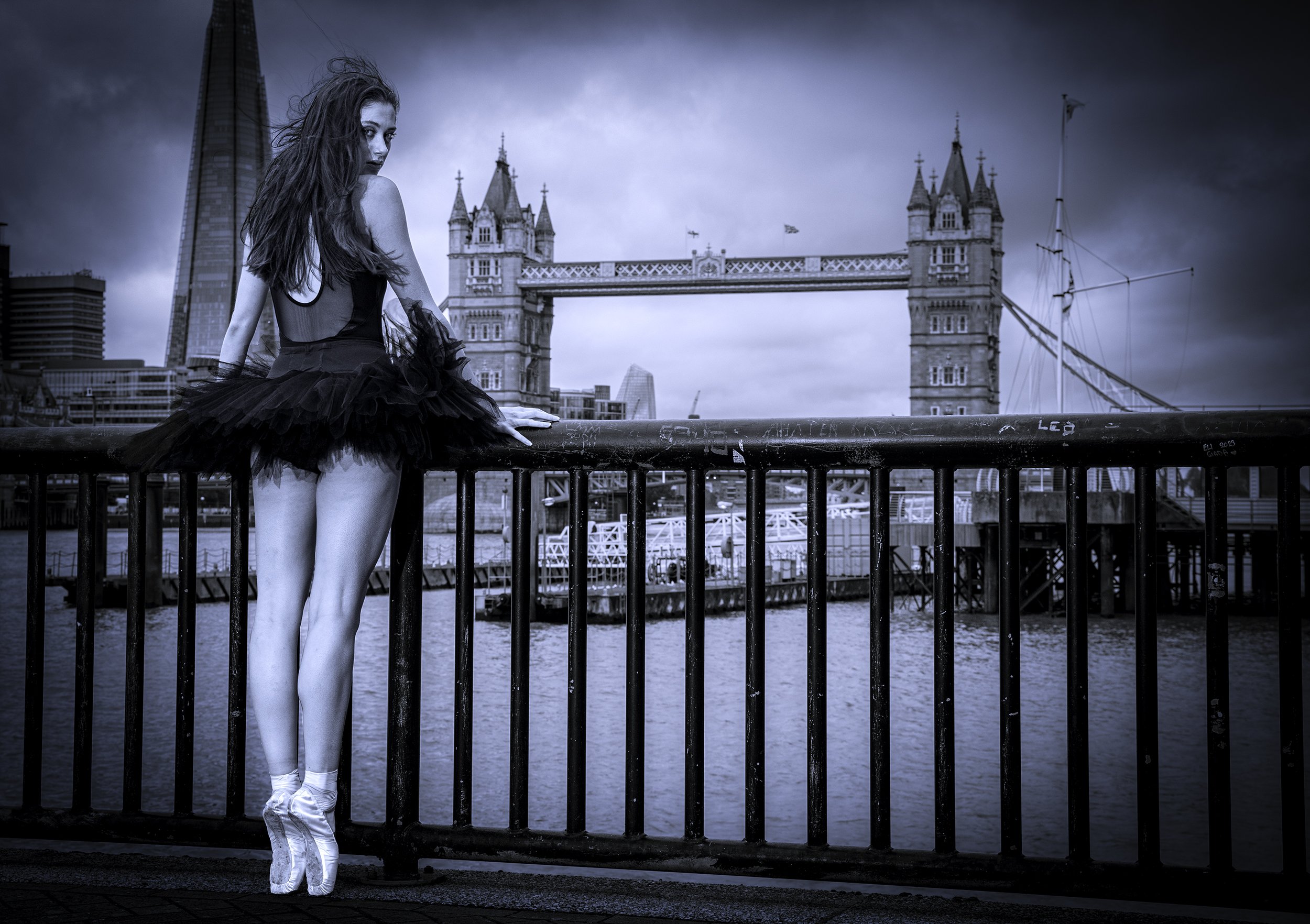 A ballet dancer dressed in black tutu and pointe shoes poses on a bridge railing with cityscape and Tower Bridge in London in the background, under a dark, cloudy sky.