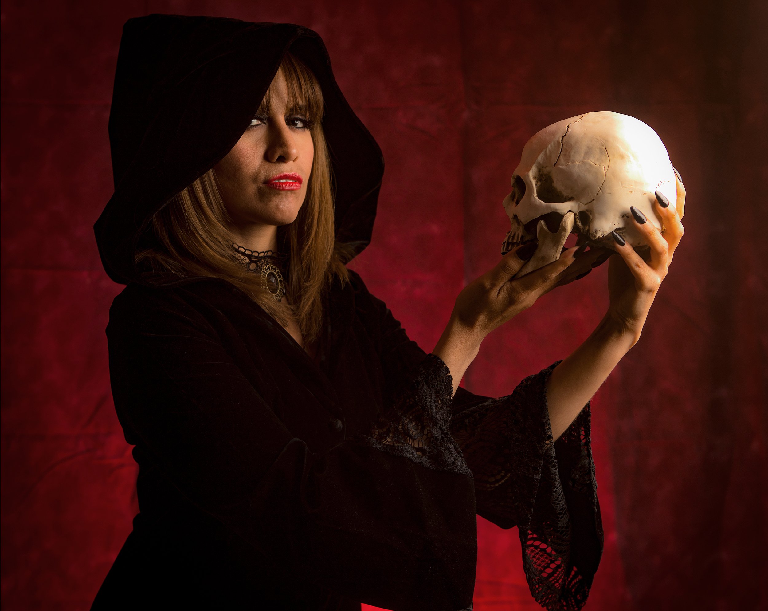 Woman dressed in black with a hood holding a human skull against a dark red background.