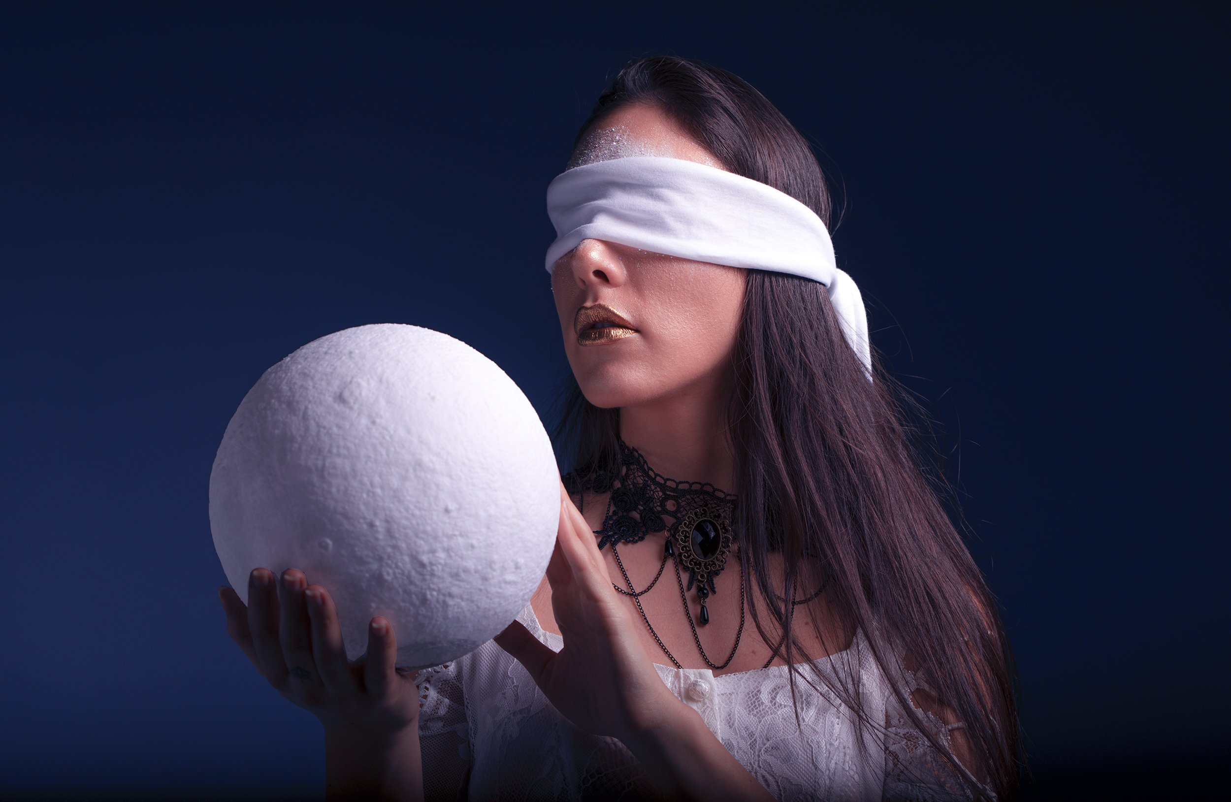 A woman with dark hair and metallic lipstick, wearing a white blindfold and gothic jewelry, holding a textured white spherical object against a dark blue background.