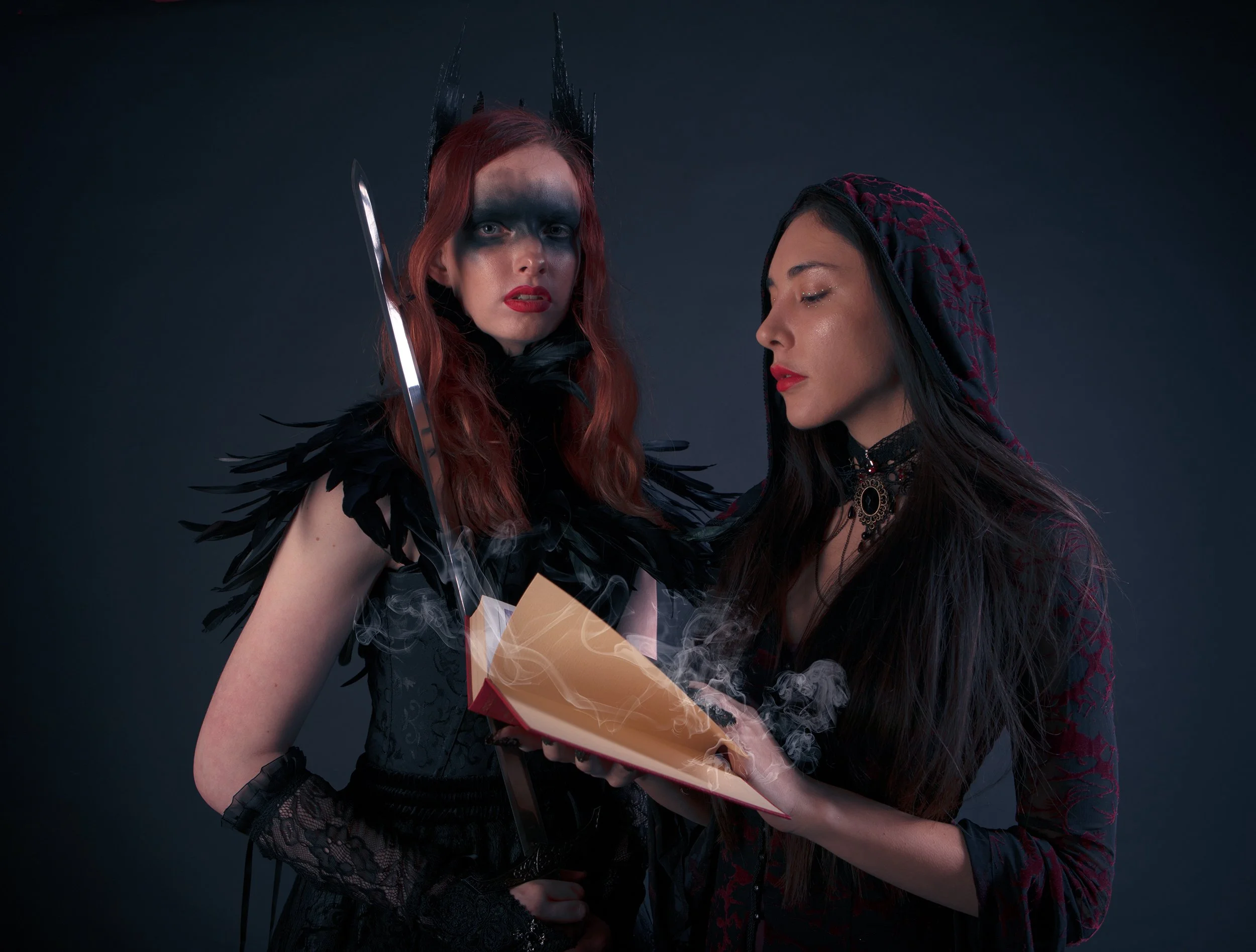 Two women dressed in dark, gothic costumes, one with red hair and the other with black hair, are reading a steaming book against a dark background.