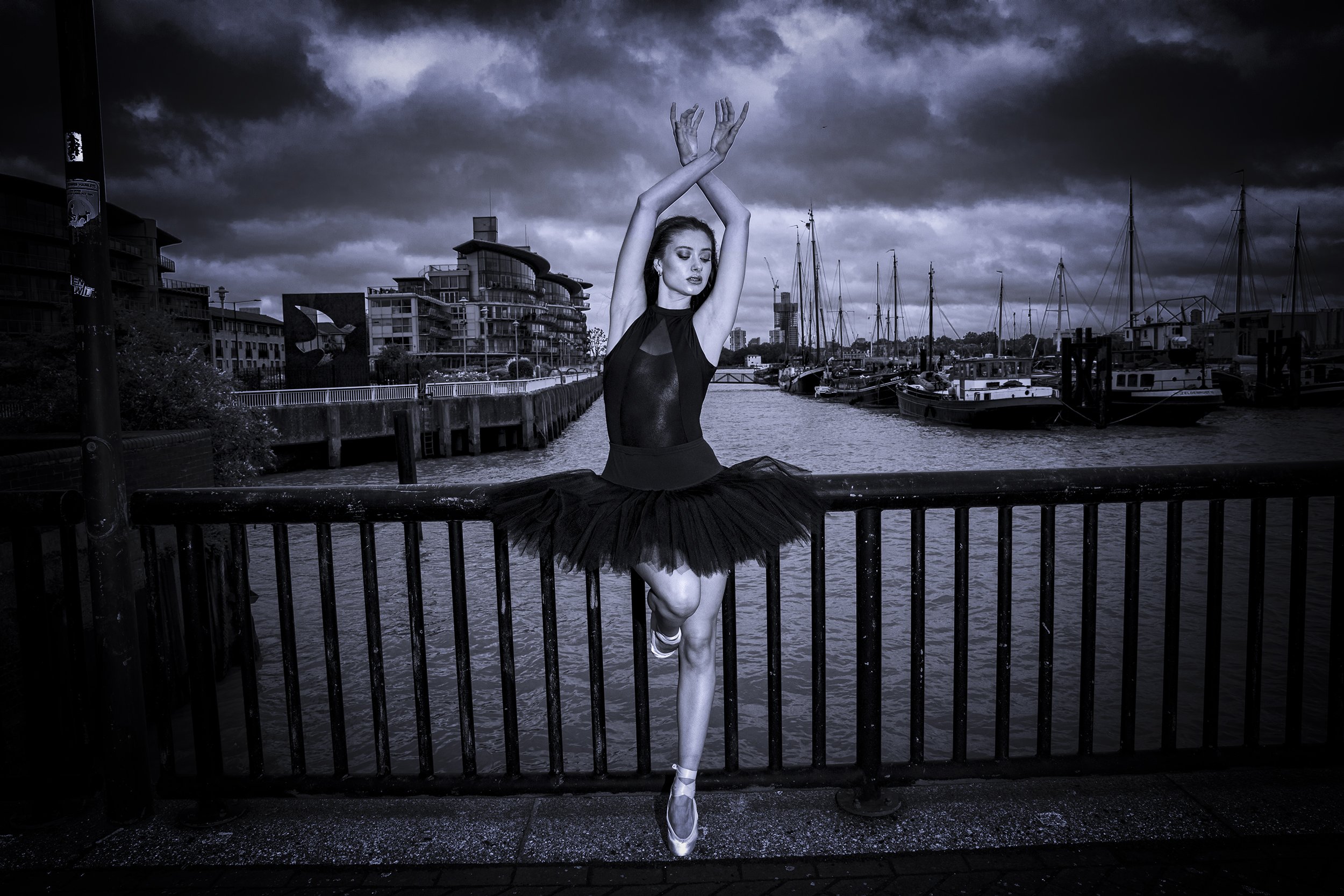 A ballerina in a black tutu and sleeveless leotard practicing ballet on a waterfront promenade with boats and modern buildings in the background under a cloudy sky.
