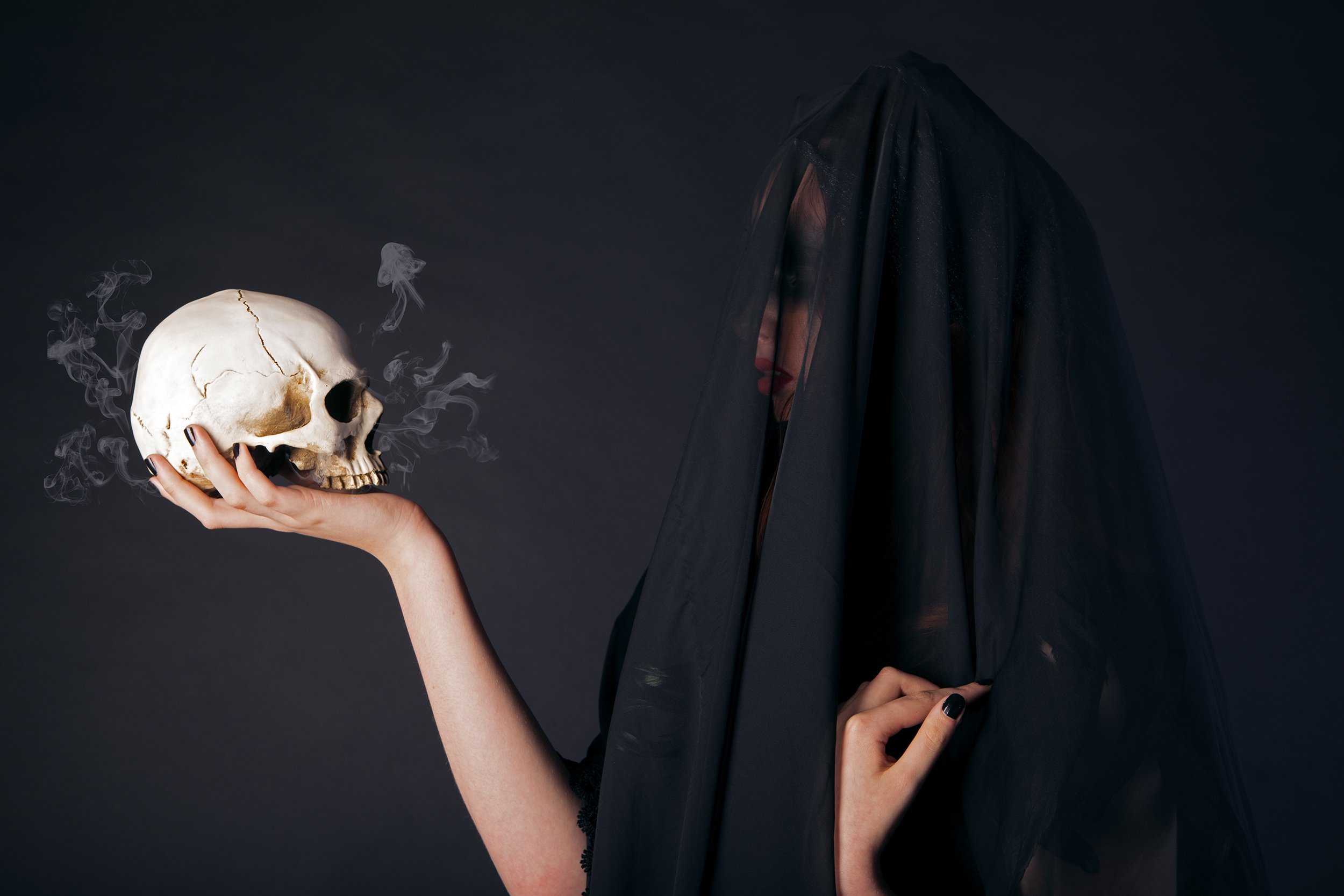 A woman dressed in black with a black veil covering her face, holding a skull emitting wisps of smoke, set against a dark background.
