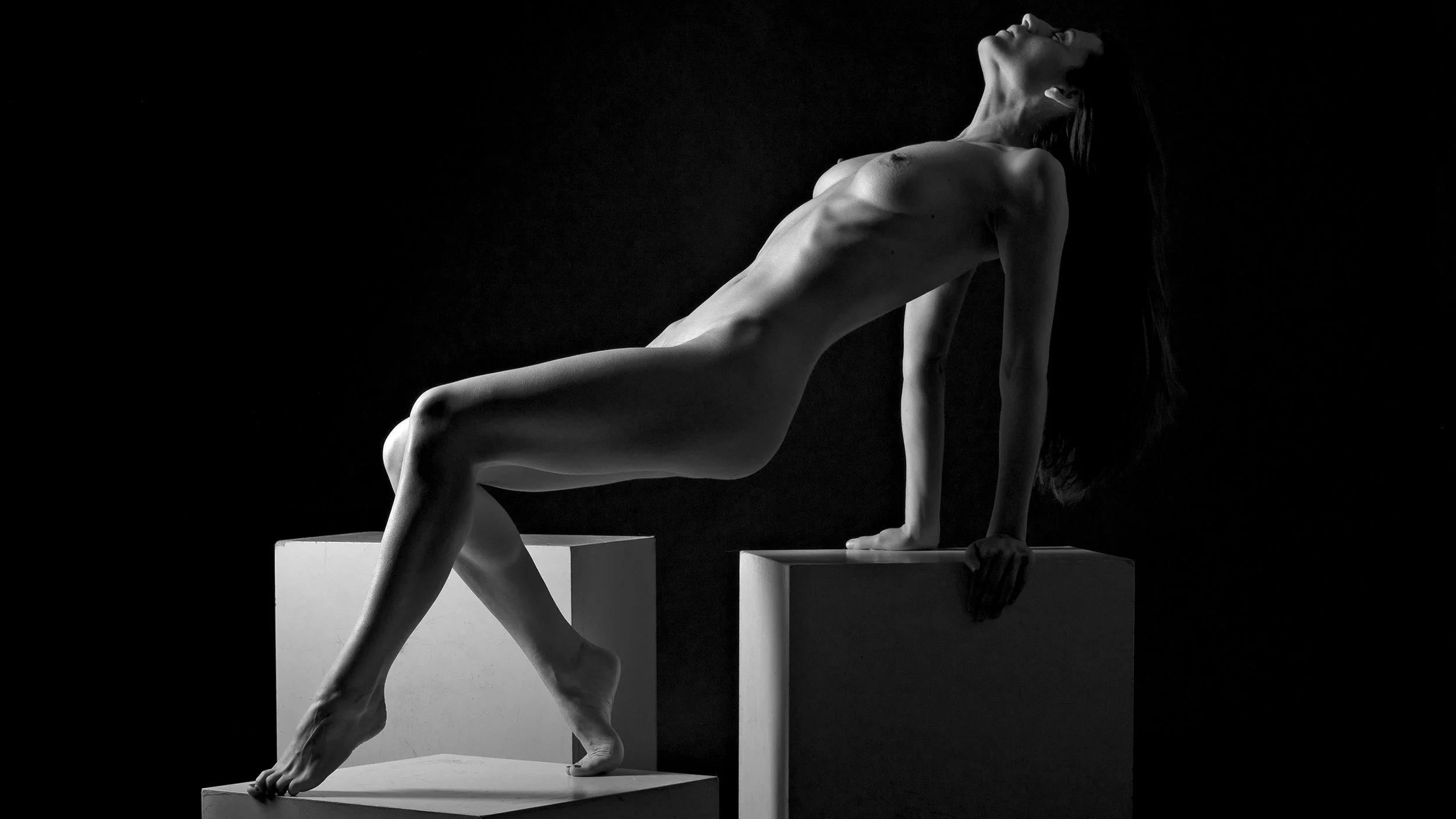 Black and white photograph of a nude woman posing on black and white cubic blocks, leaning back with her head tilted upward and her arms supporting her on the blocks.