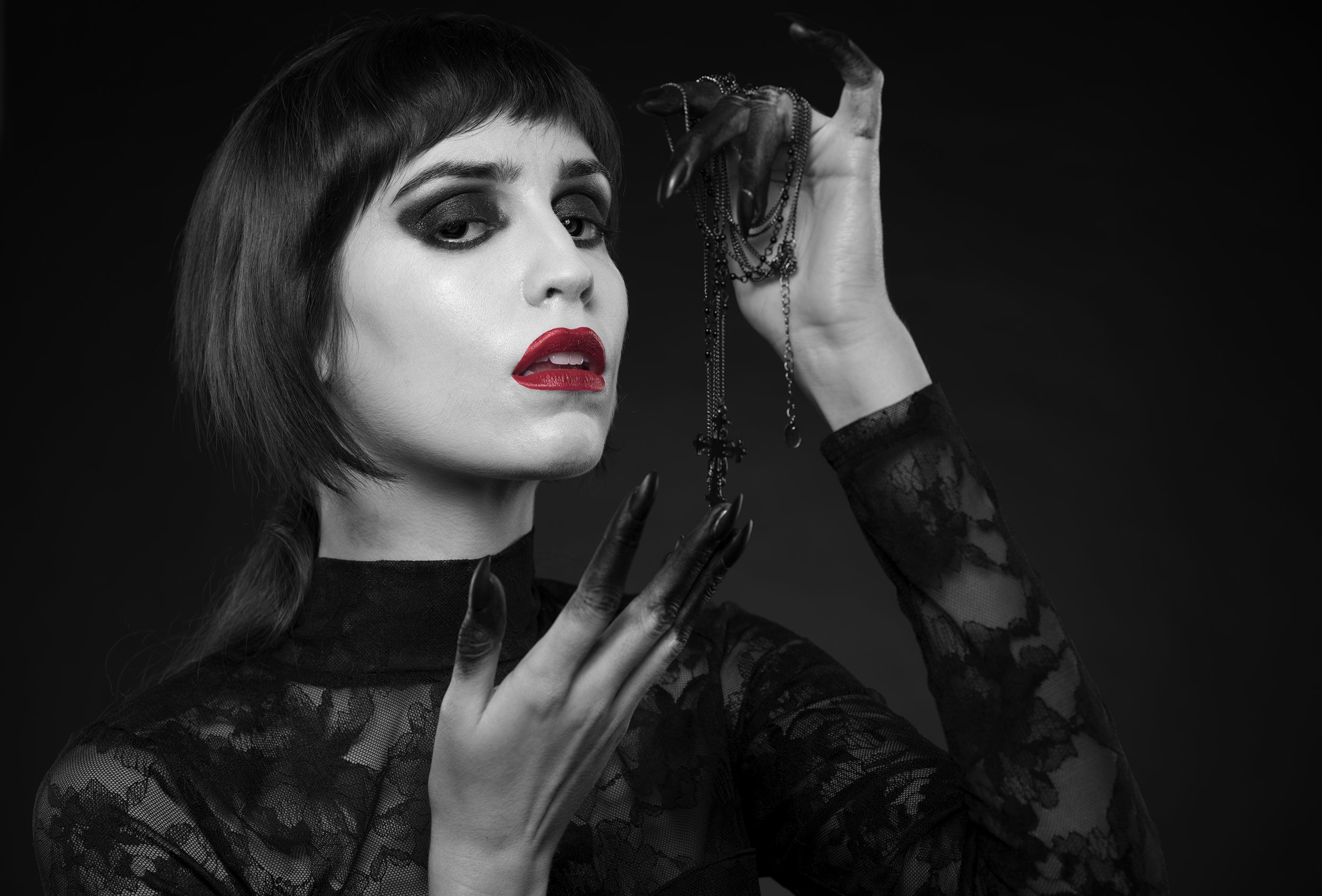 A woman with short dark hair and in gothic style, wearing black lace clothing, holds black rosary beads with a cross in one hand, with a serious expression, against a dark background.
