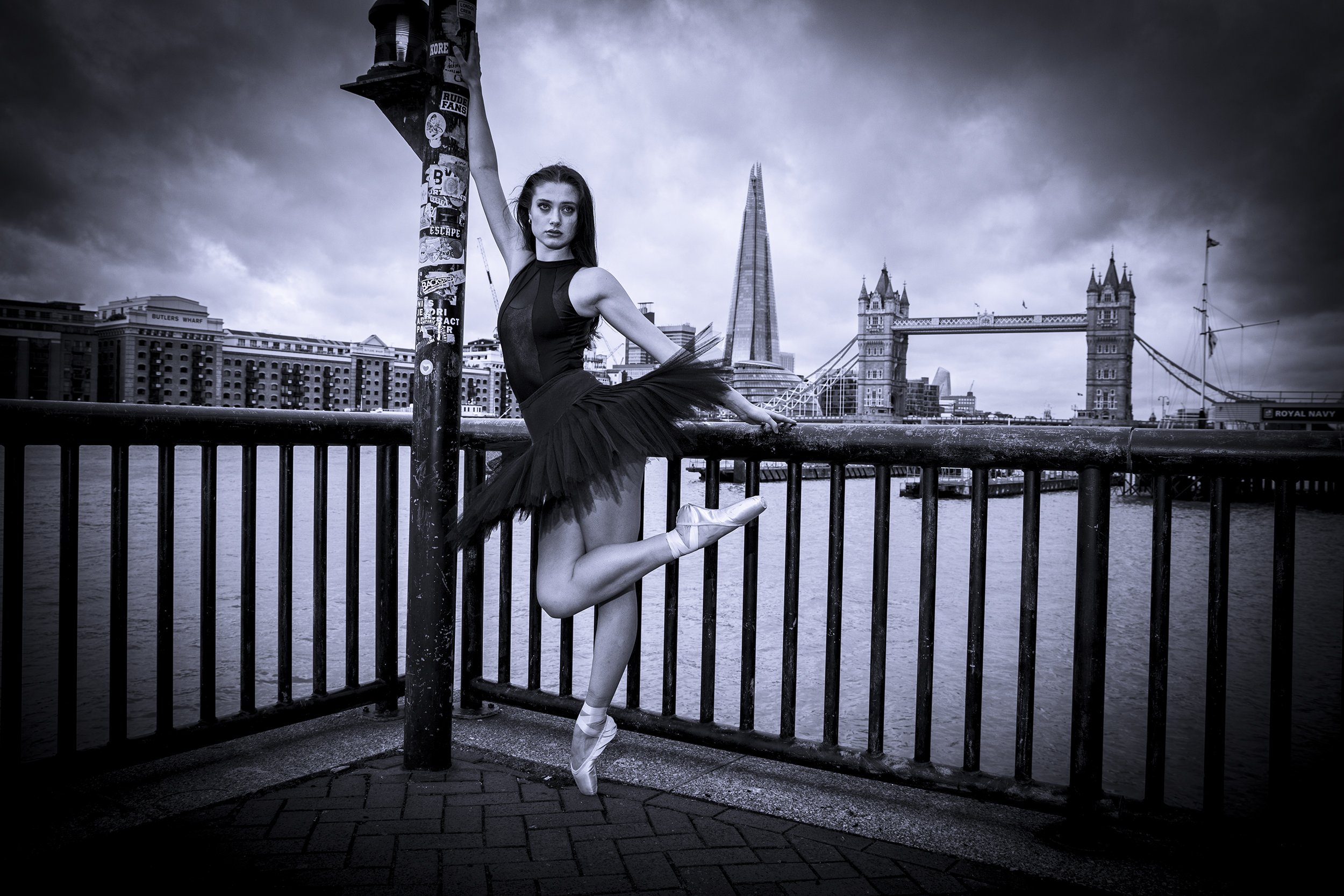A ballet dancer in a tutu and pointe shoes posing on a riverside promenade with the Tower Bridge and Tower of London in the background, in black and white.