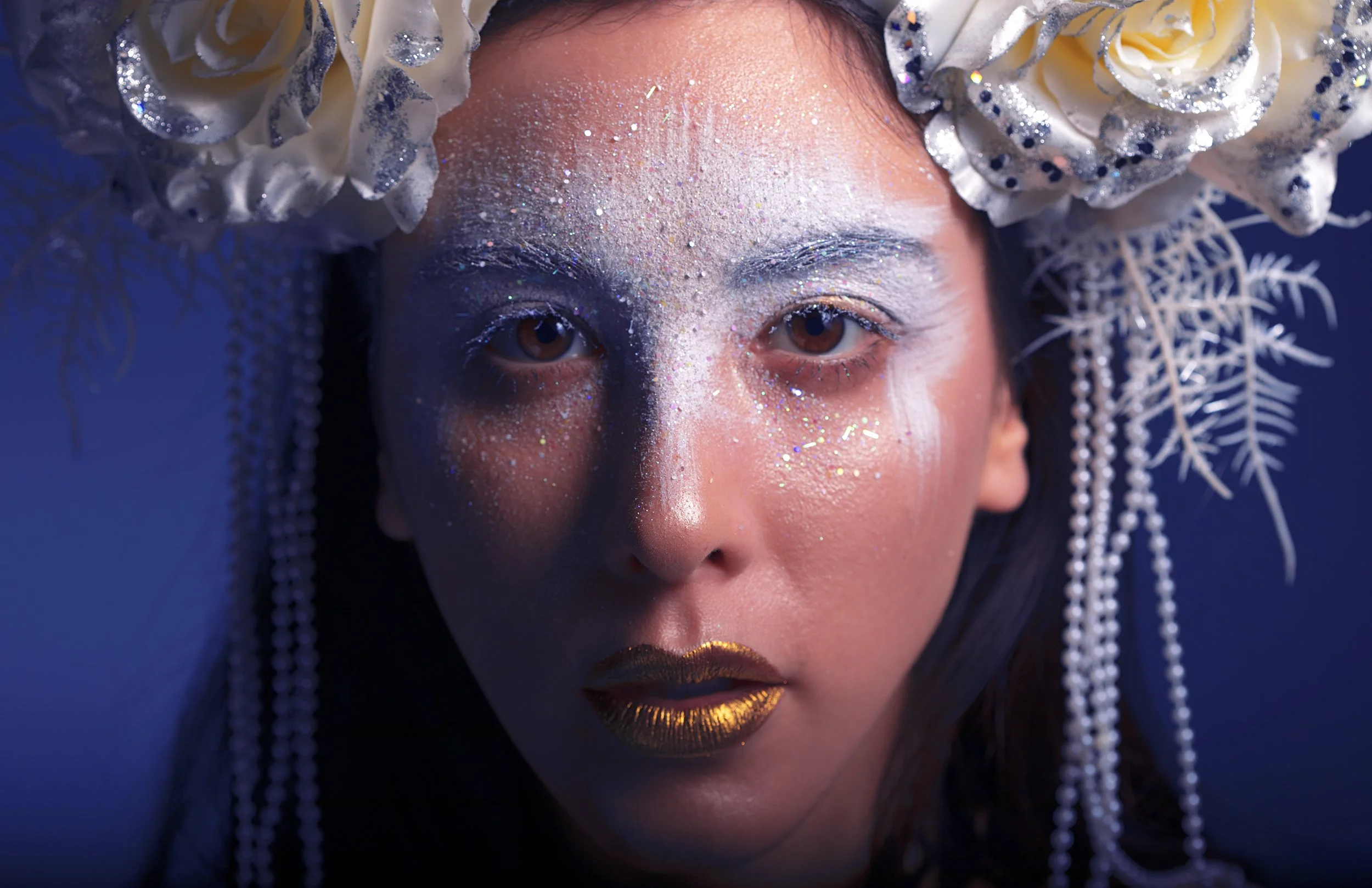 Close-up of a woman's face with glitter and makeup, wearing a headdress decorated with white flowers and dangling beads, against a dark background.